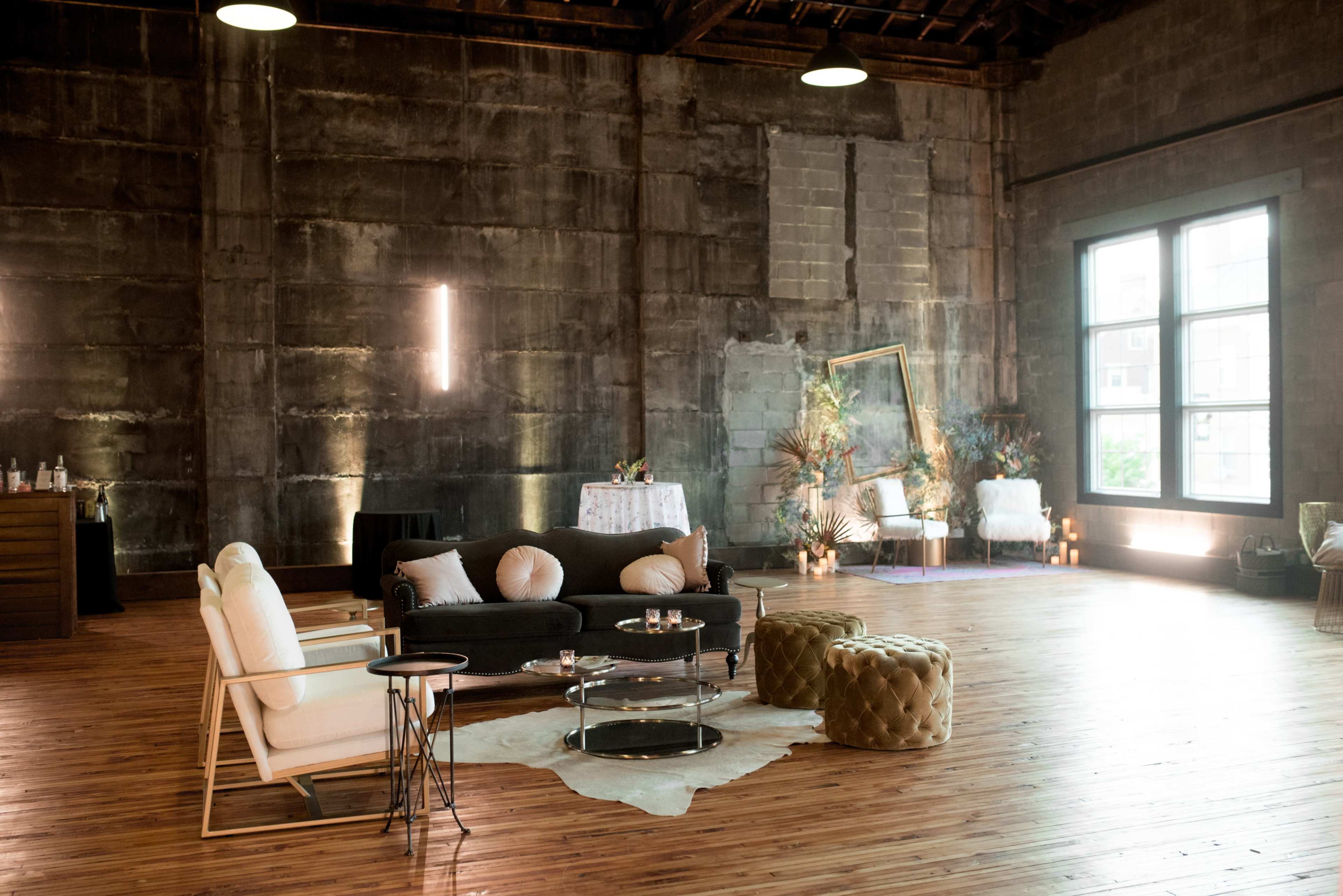 A spacious interior with wooden floors, featuring a black sofa, a white armchair, round ottomans, and decorative plants in a well-lit setting.
