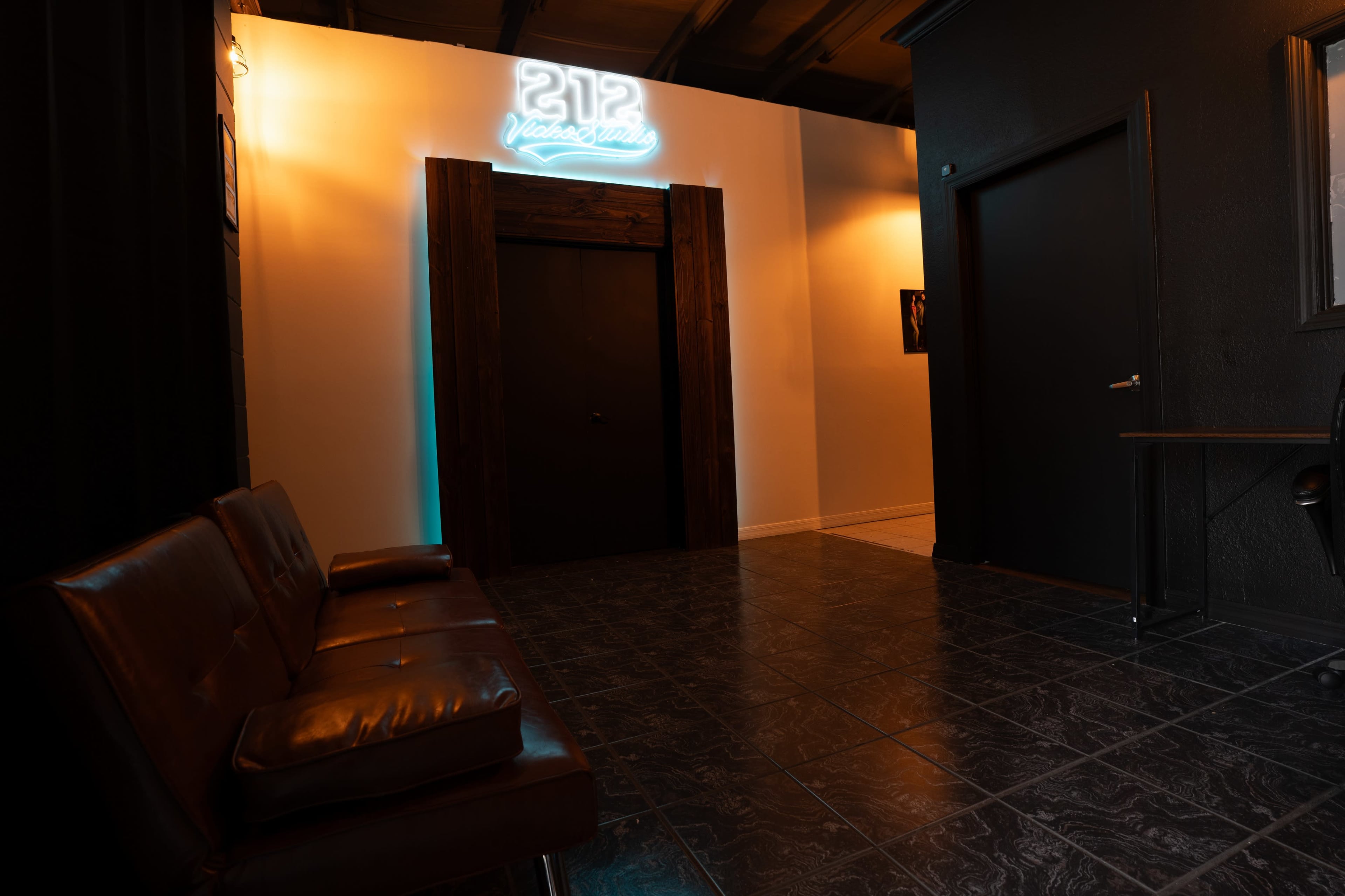 The image shows a dimly lit hallway featuring a brown leather couch, a door framed by wooden accents, and a neon sign reading "212 Photography."