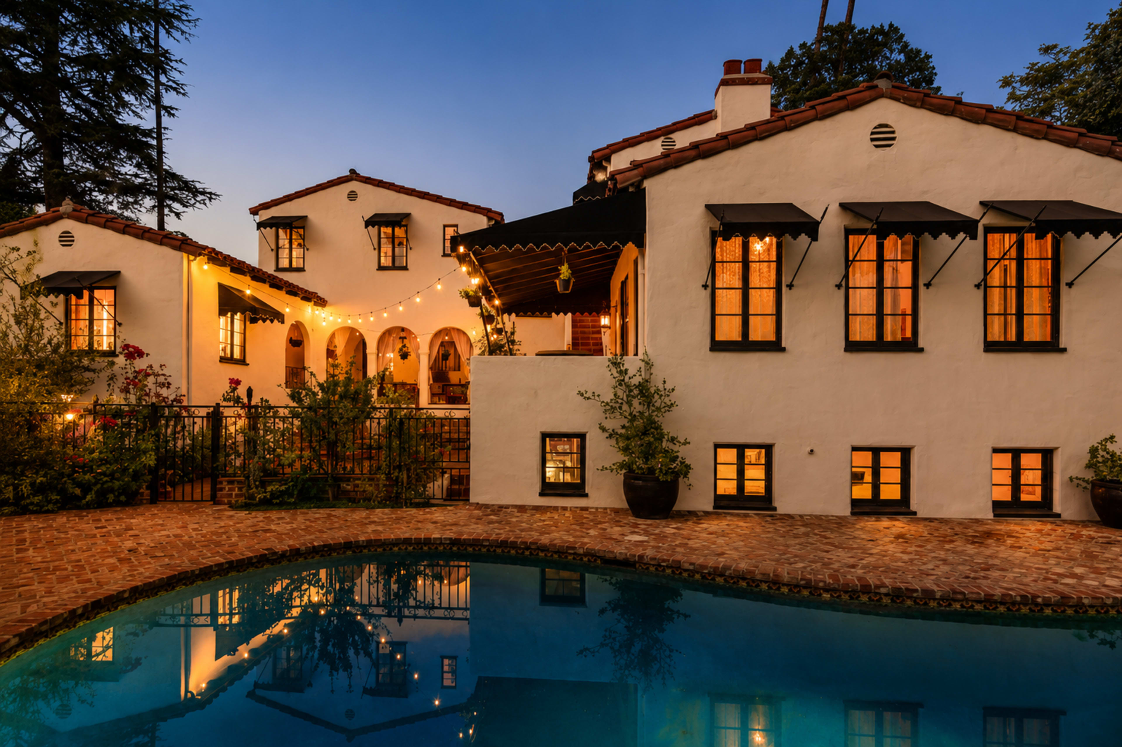 A warm-lit, Mediterranean-style home is reflected in a swimming pool at dusk, with string lights adorning the exterior.