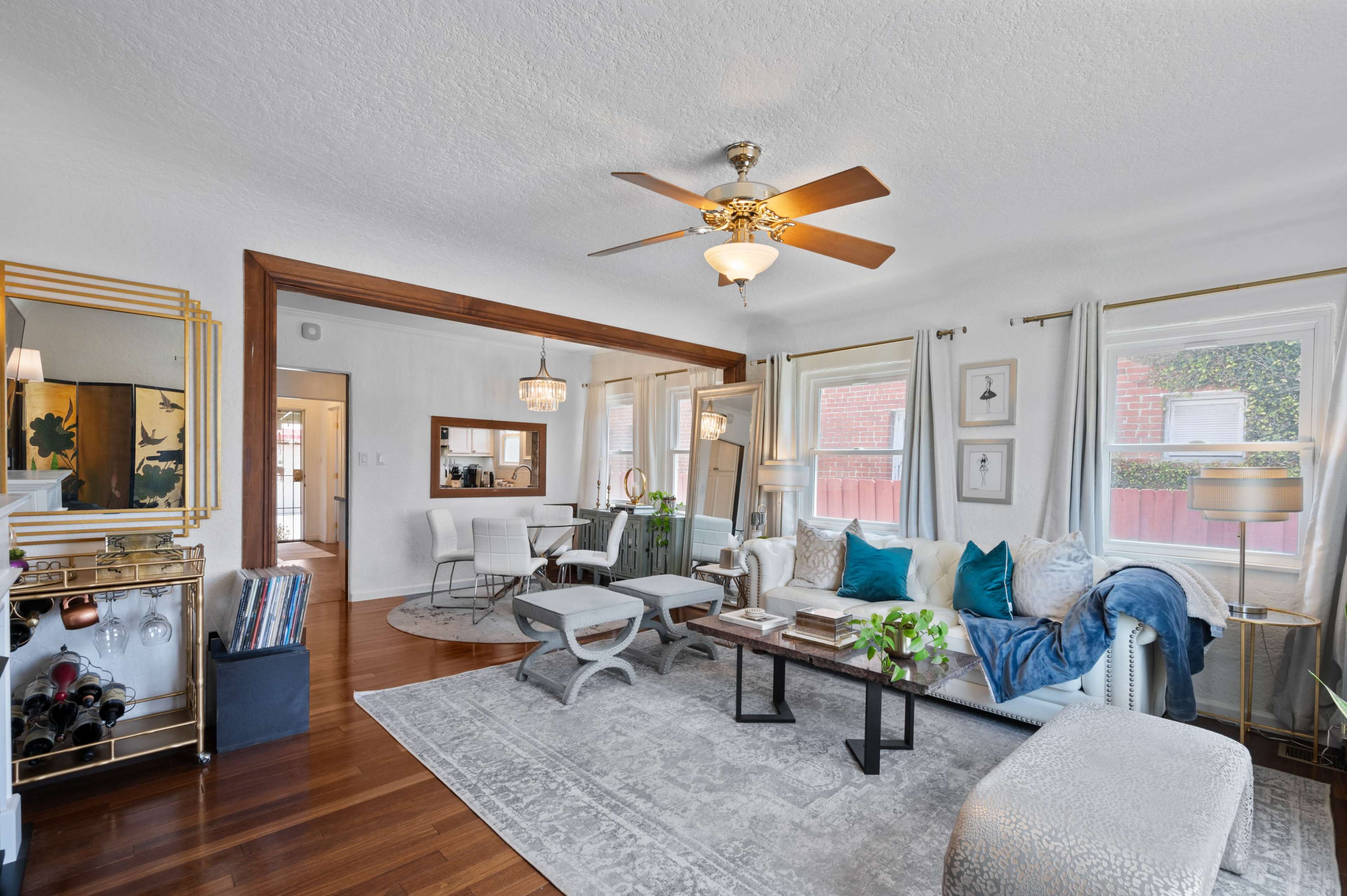 A spacious living room features a seating area with a cream-colored sofa, a coffee table, and a dining nook, all illuminated by natural light from large windows.