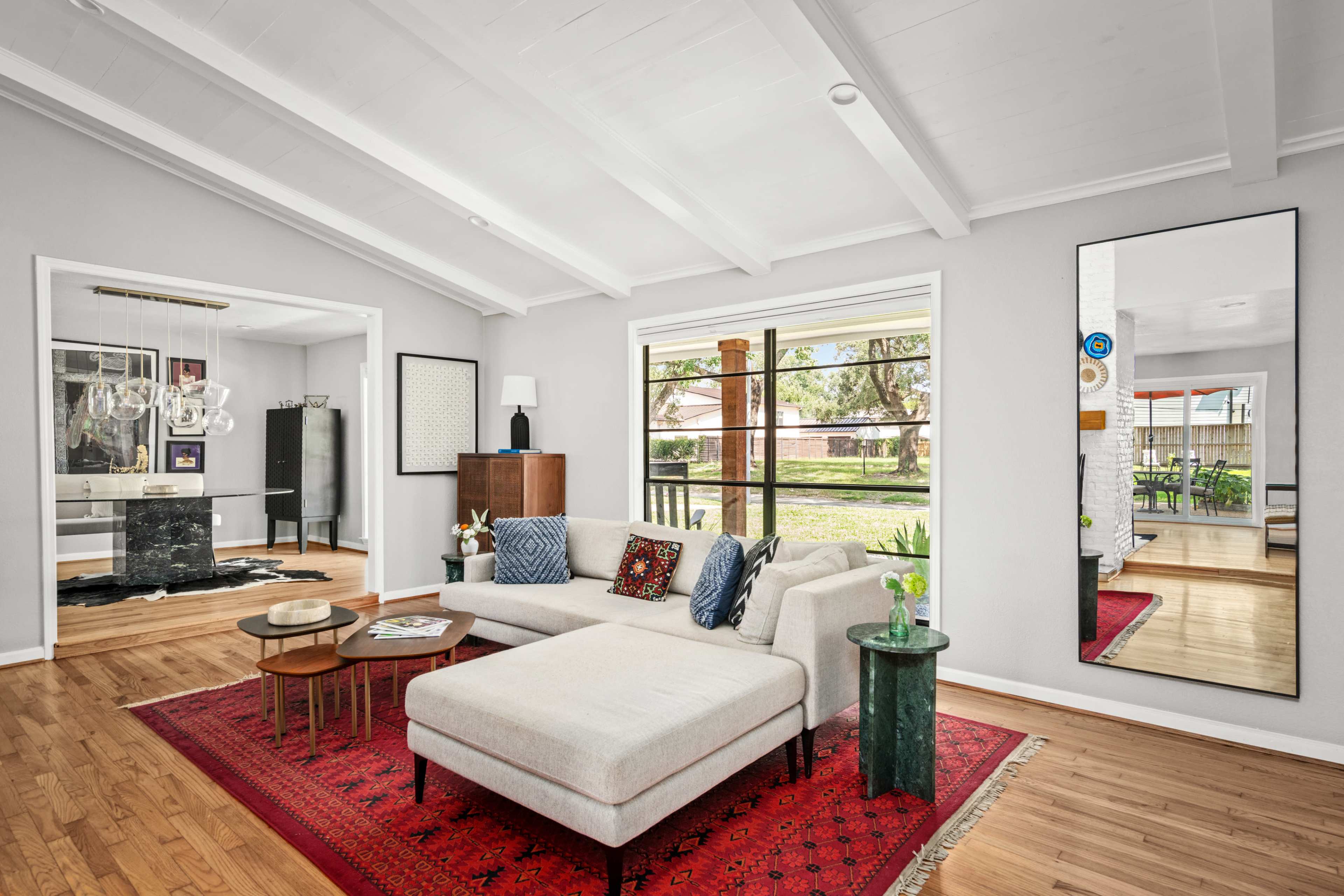 The image shows a modern living room with a light-colored sectional sofa, a red patterned area rug, and large windows that provide a view of a backyard.