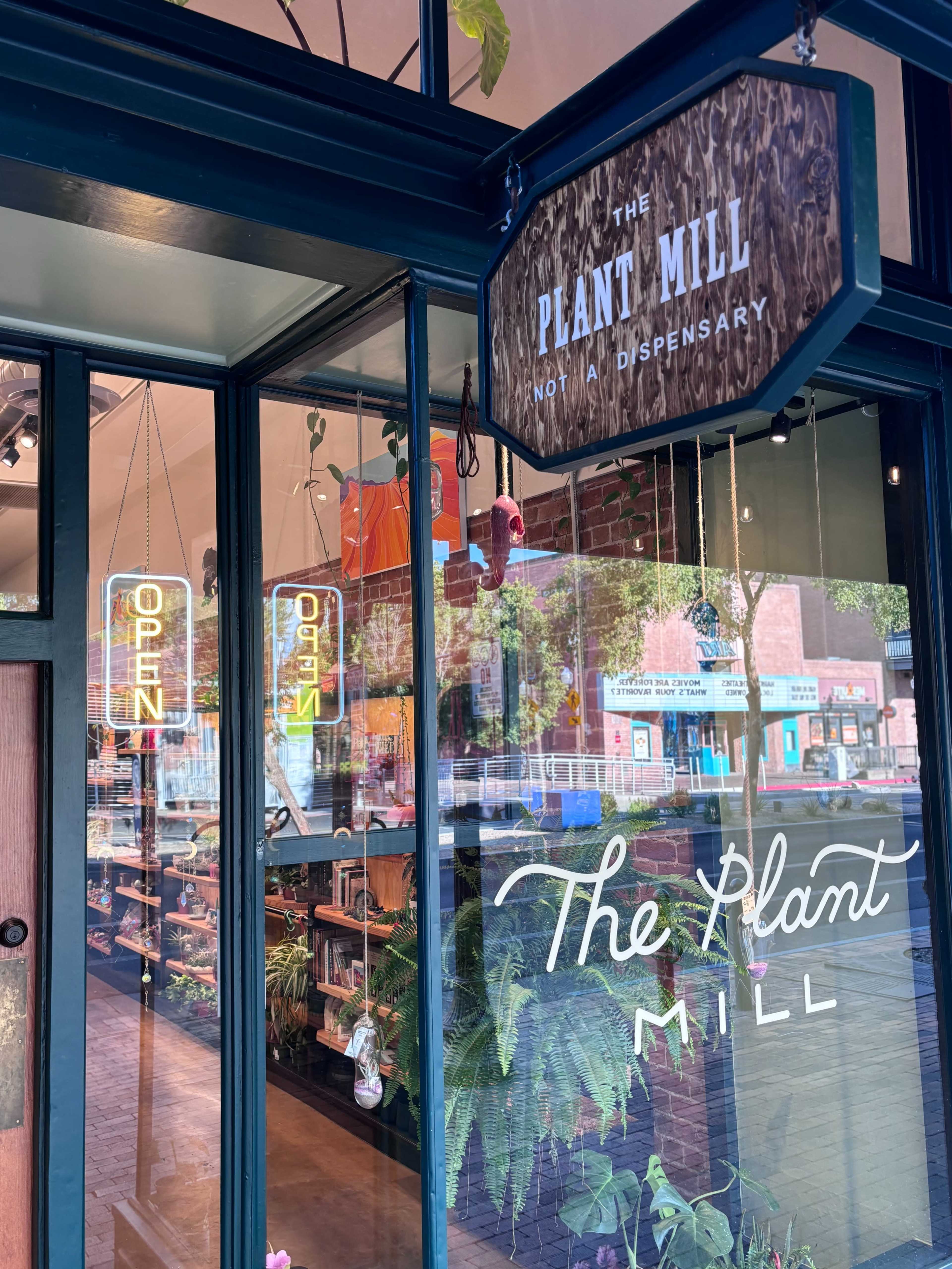 The image shows the entrance of a plant shop called "The Plant Mill," featuring a wooden sign and neon "OPEN" signs, with shelves of plants visible inside.