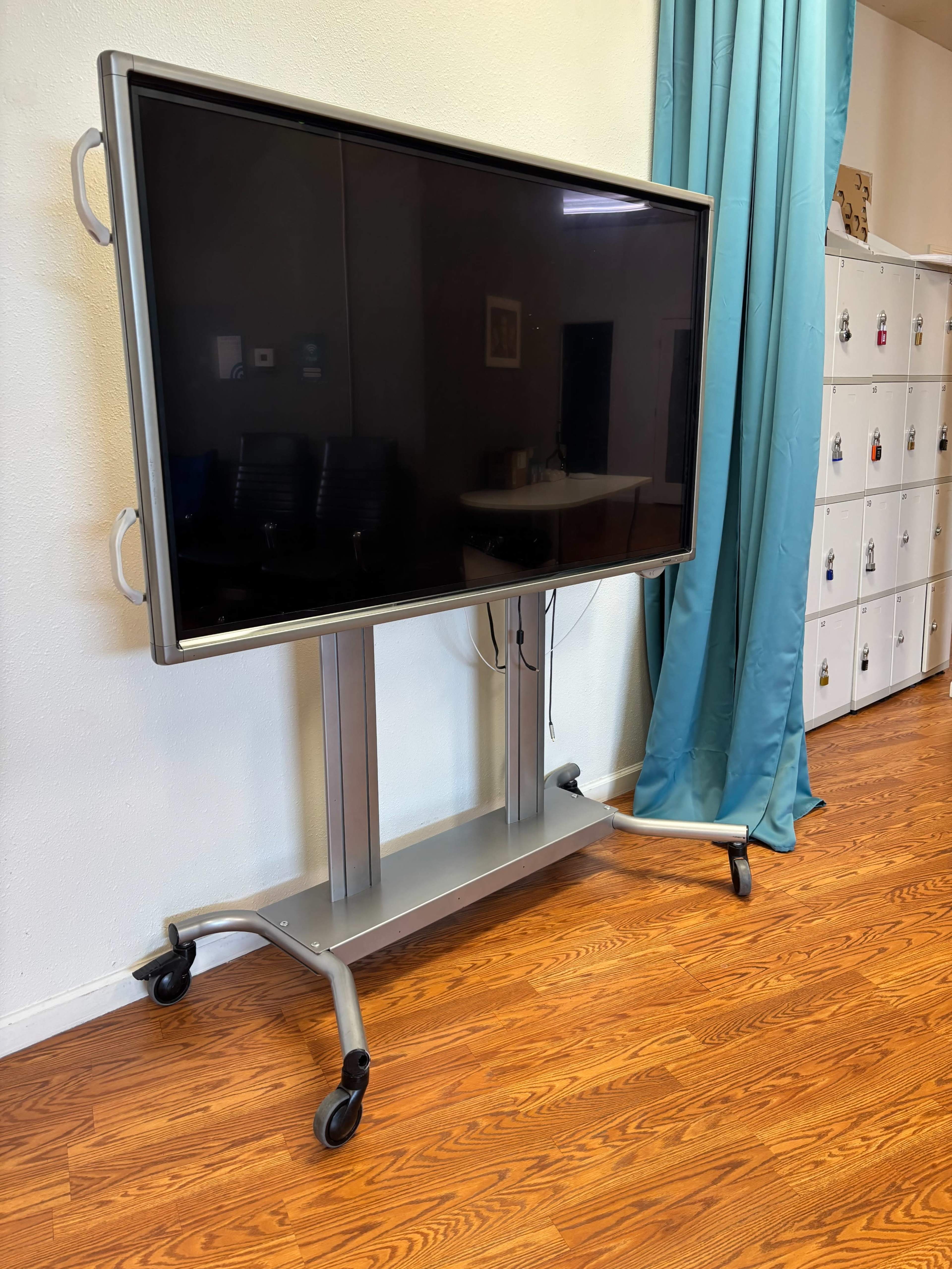 A large television mounted on a wheeled mobile stand is positioned near a curtain and storage lockers in a room with wooden flooring.