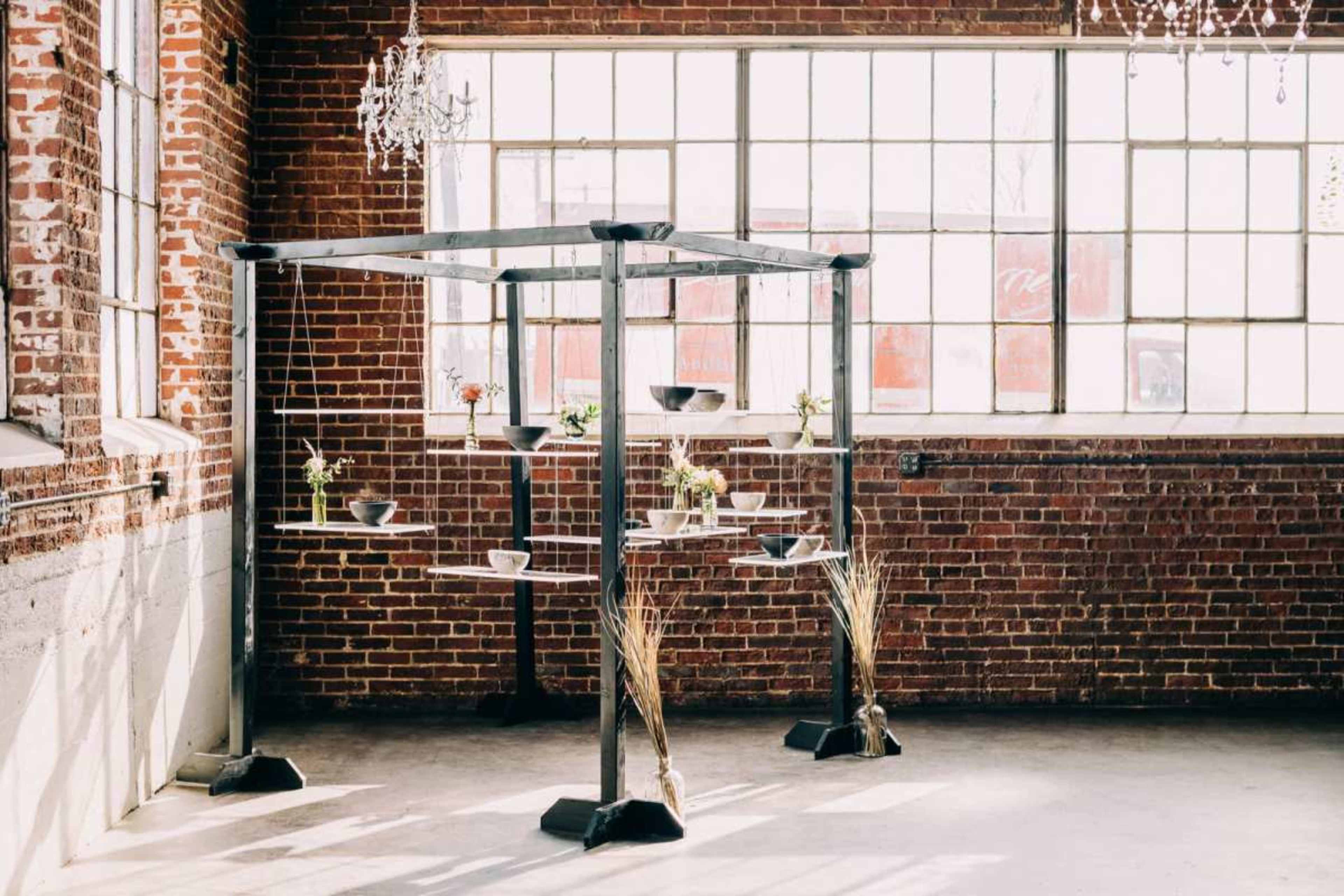 A metal display structure with shelves is filled with potted plants, positioned near large windows in a rustic brick interior.