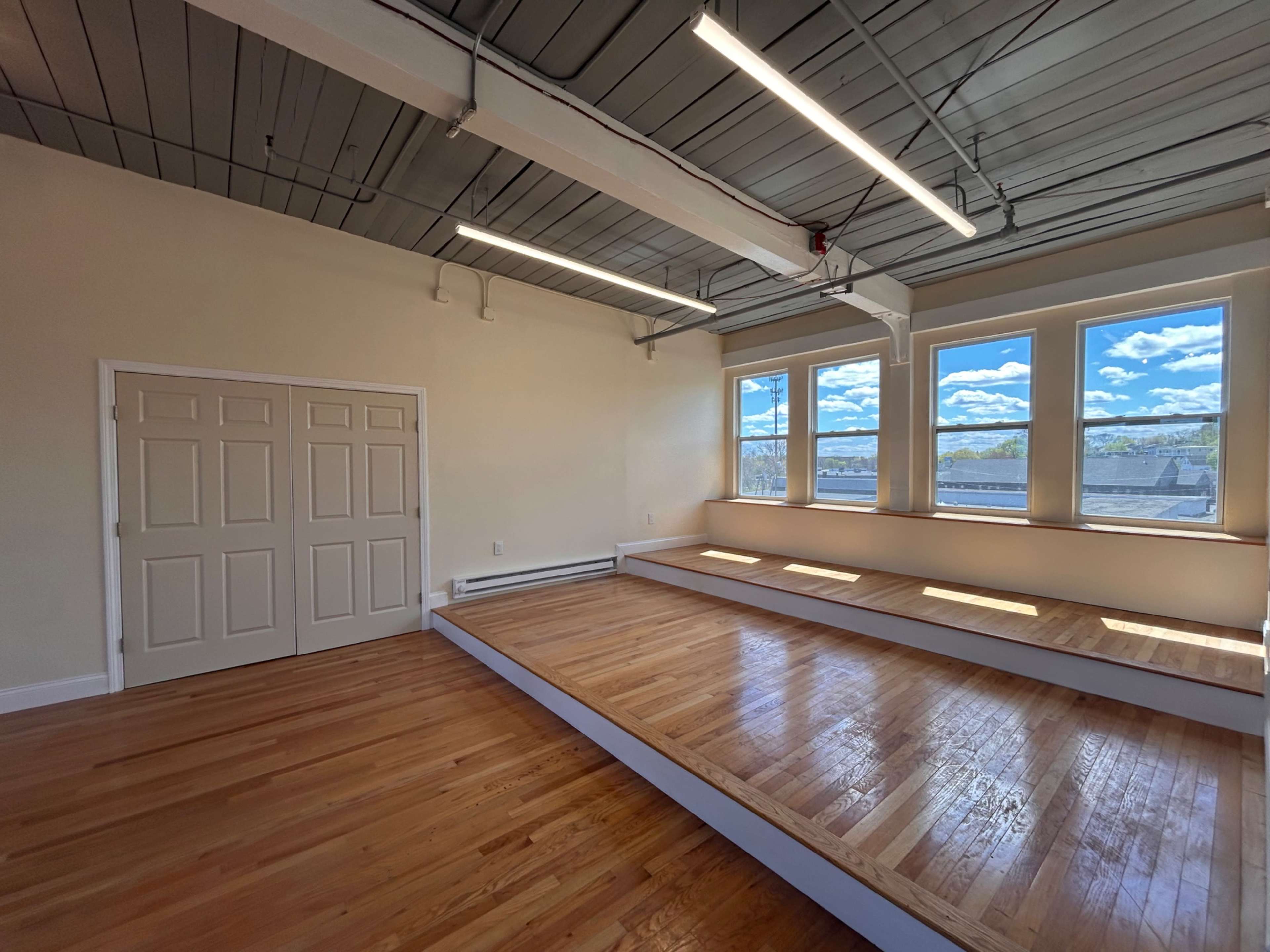 The image shows an empty room with hardwood flooring, two large windows, and a raised platform along one wall.