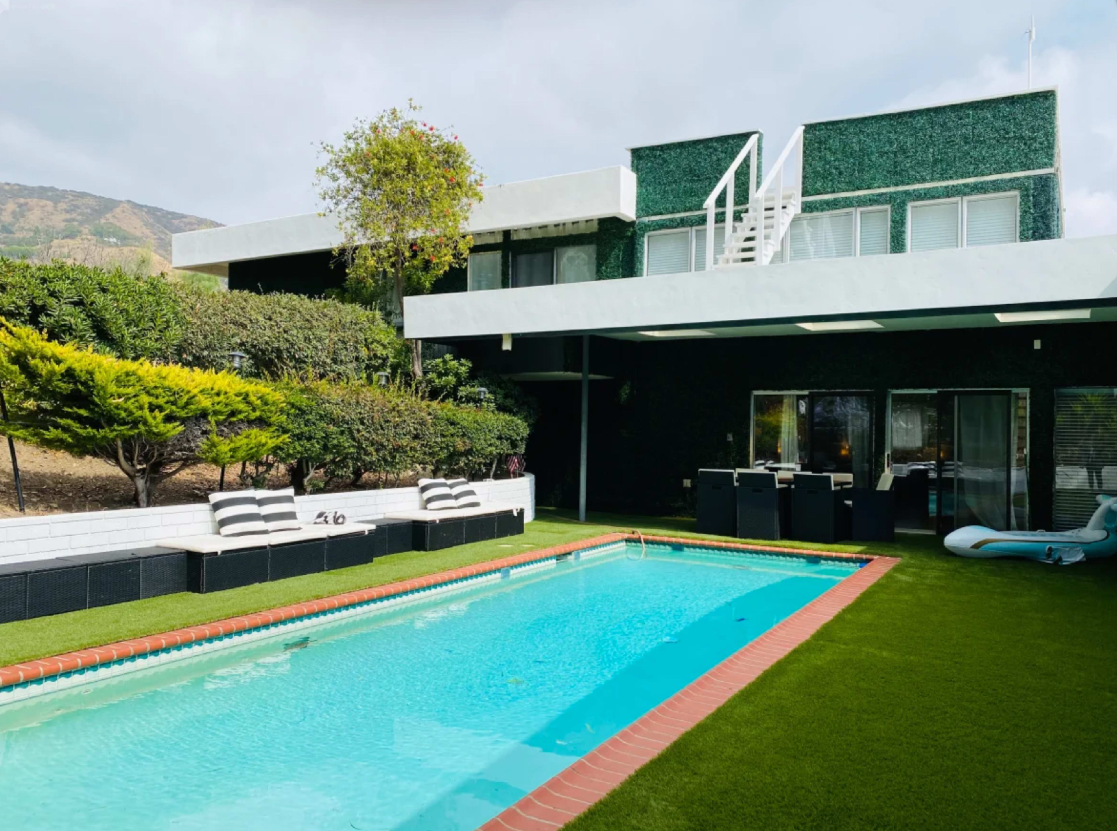 A modern house with a green exterior, a swimming pool, and a landscaped yard featuring seating areas.