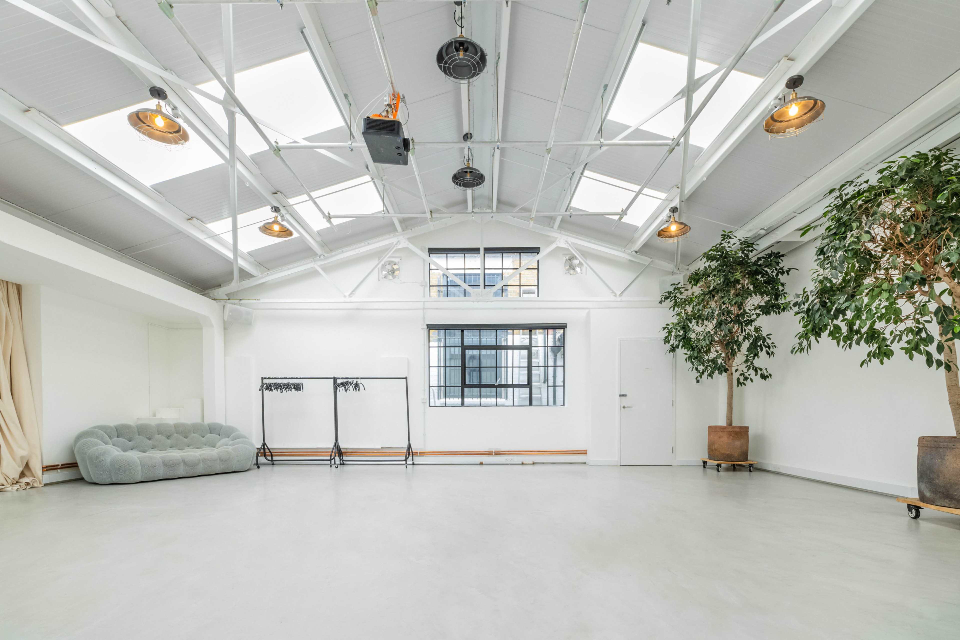 A spacious, well-lit room with a high ceiling, featuring large windows, a gray couch, a clothing rack, and potted plants.