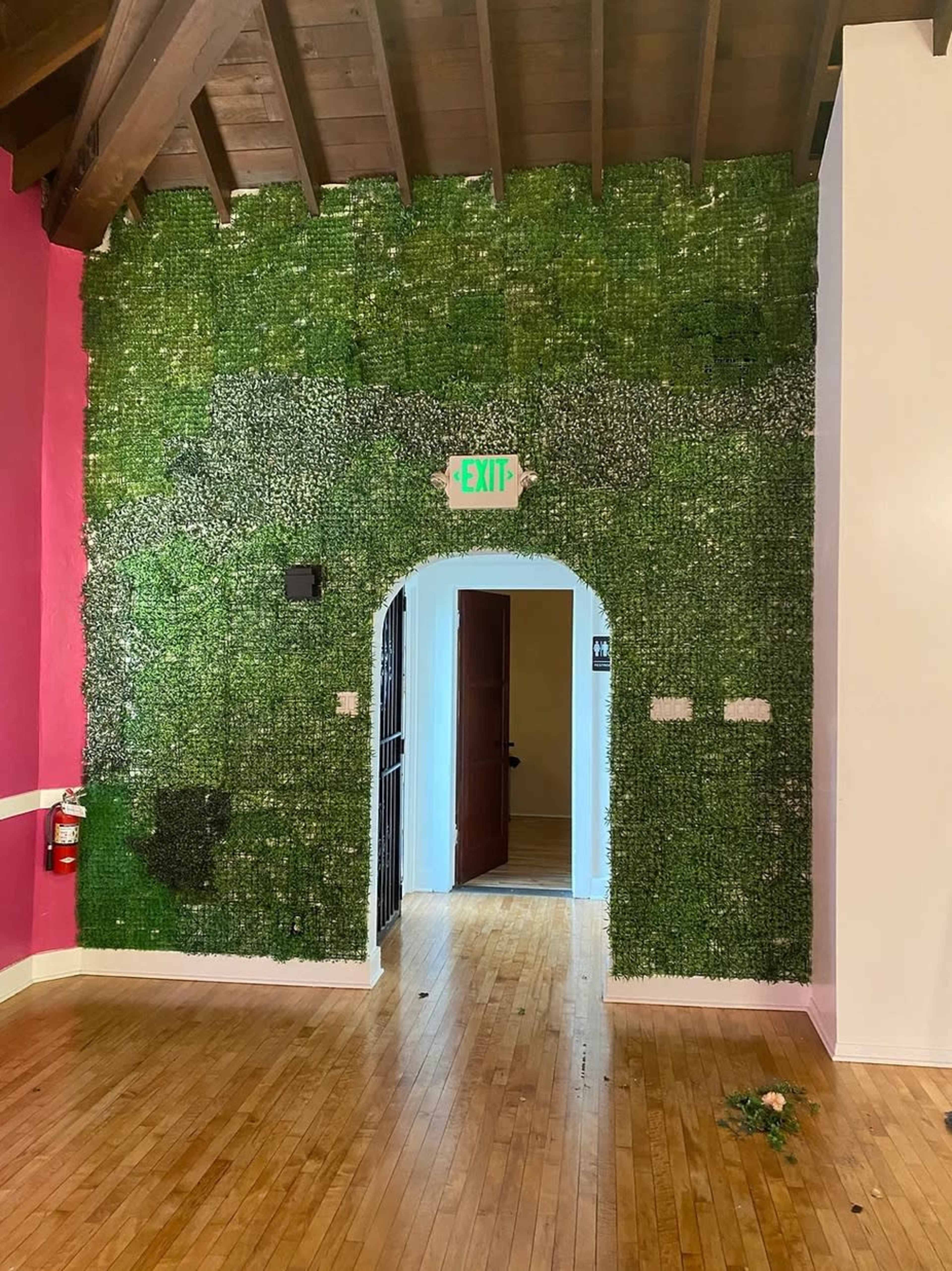 A wall covered with artificial greenery features an archway leading to a doorway, with a fire extinguisher and an exit sign visible.