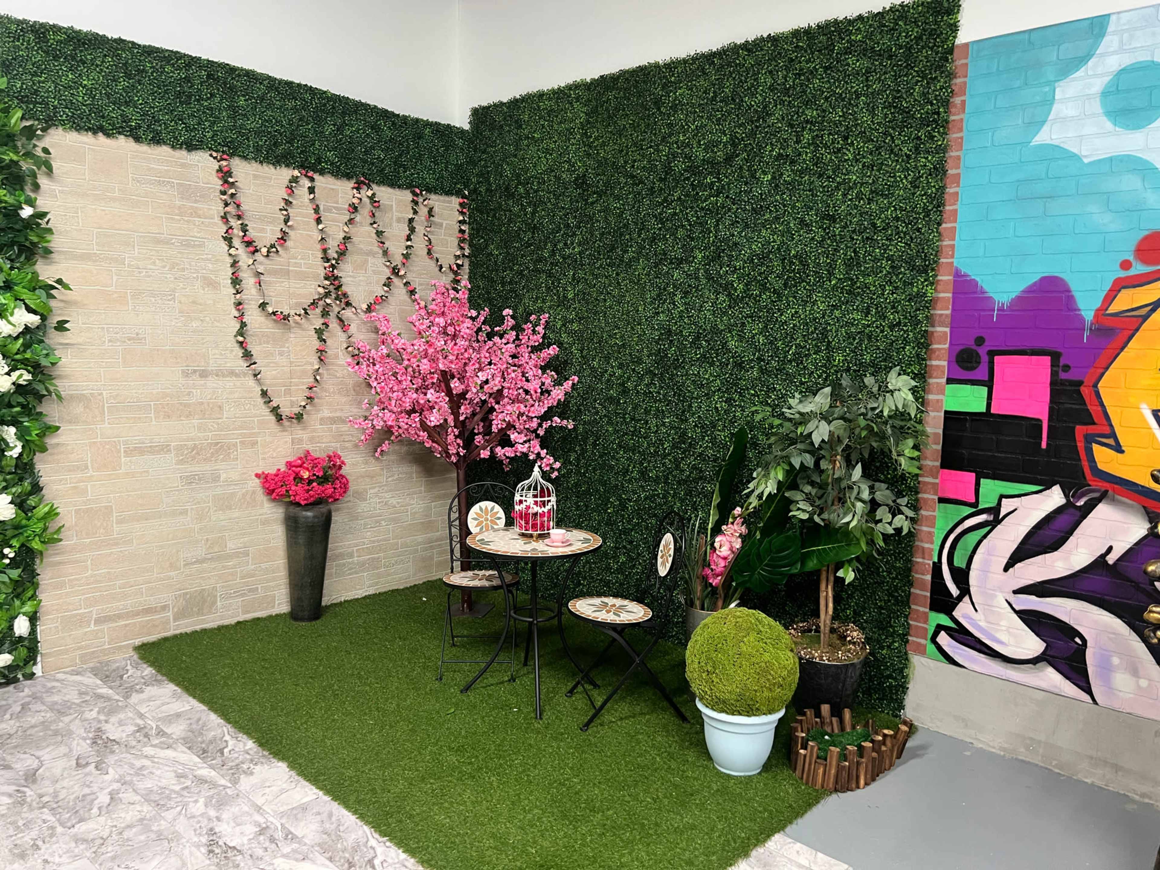 The image shows a decorative corner featuring artificial greenery, a small round table with chairs, and ornamental plants, including pink flowers and a moss-covered sphere.