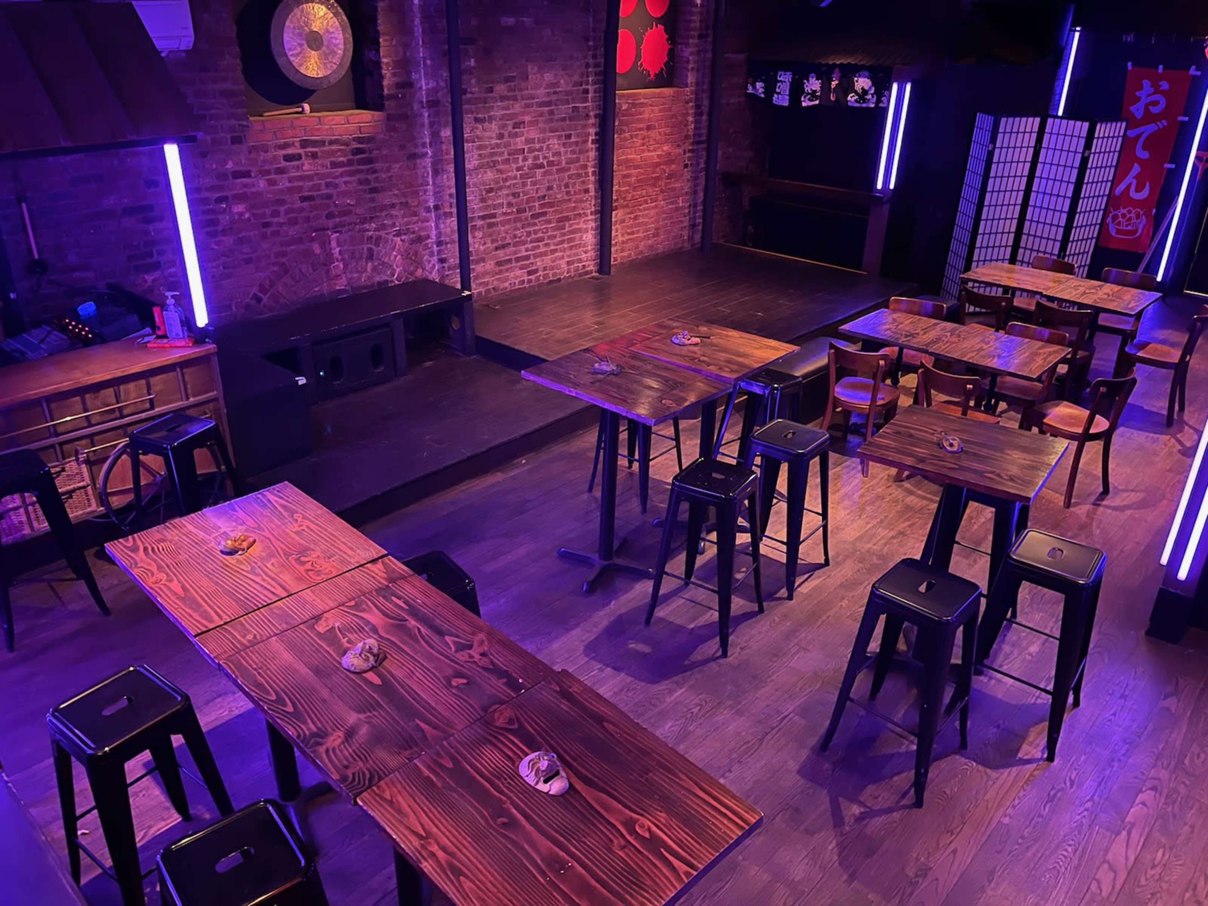 The image shows a dimly lit venue with wooden tables and stools arranged around a small stage, set against a brick wall with neon lighting.