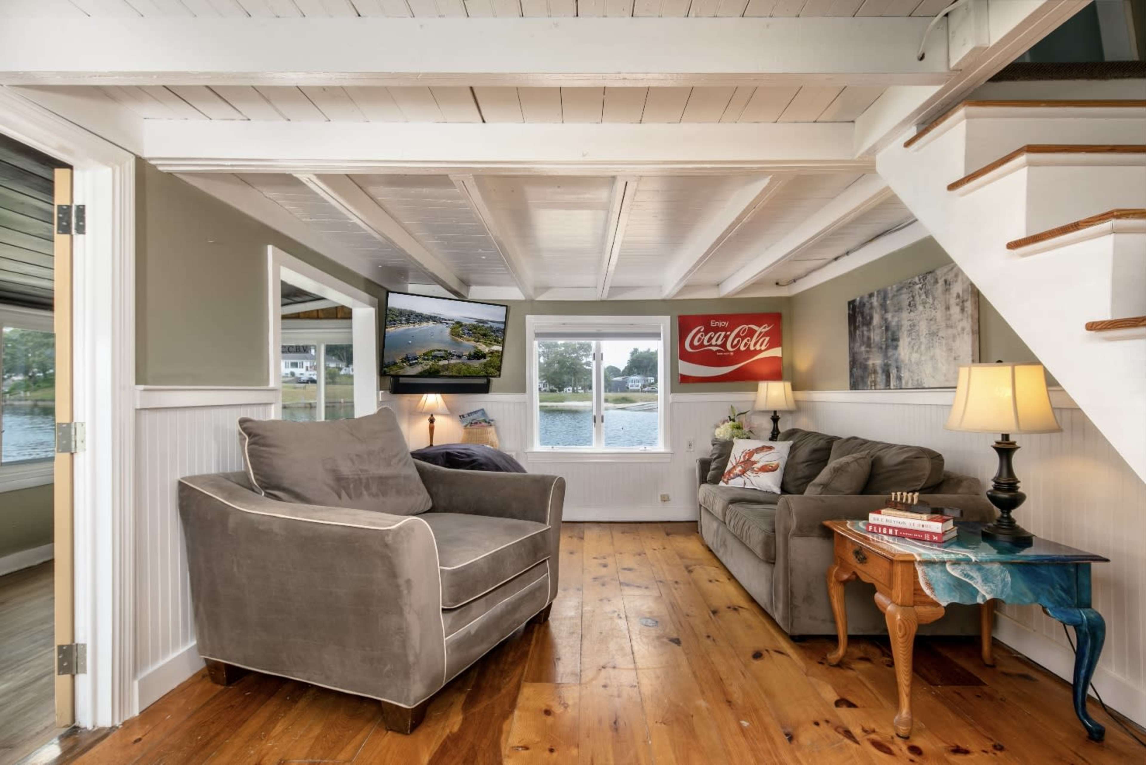 A cozy living room with a gray armchair and couch, a wooden floor, wall-mounted TV, and a staircase leading to an upper level.