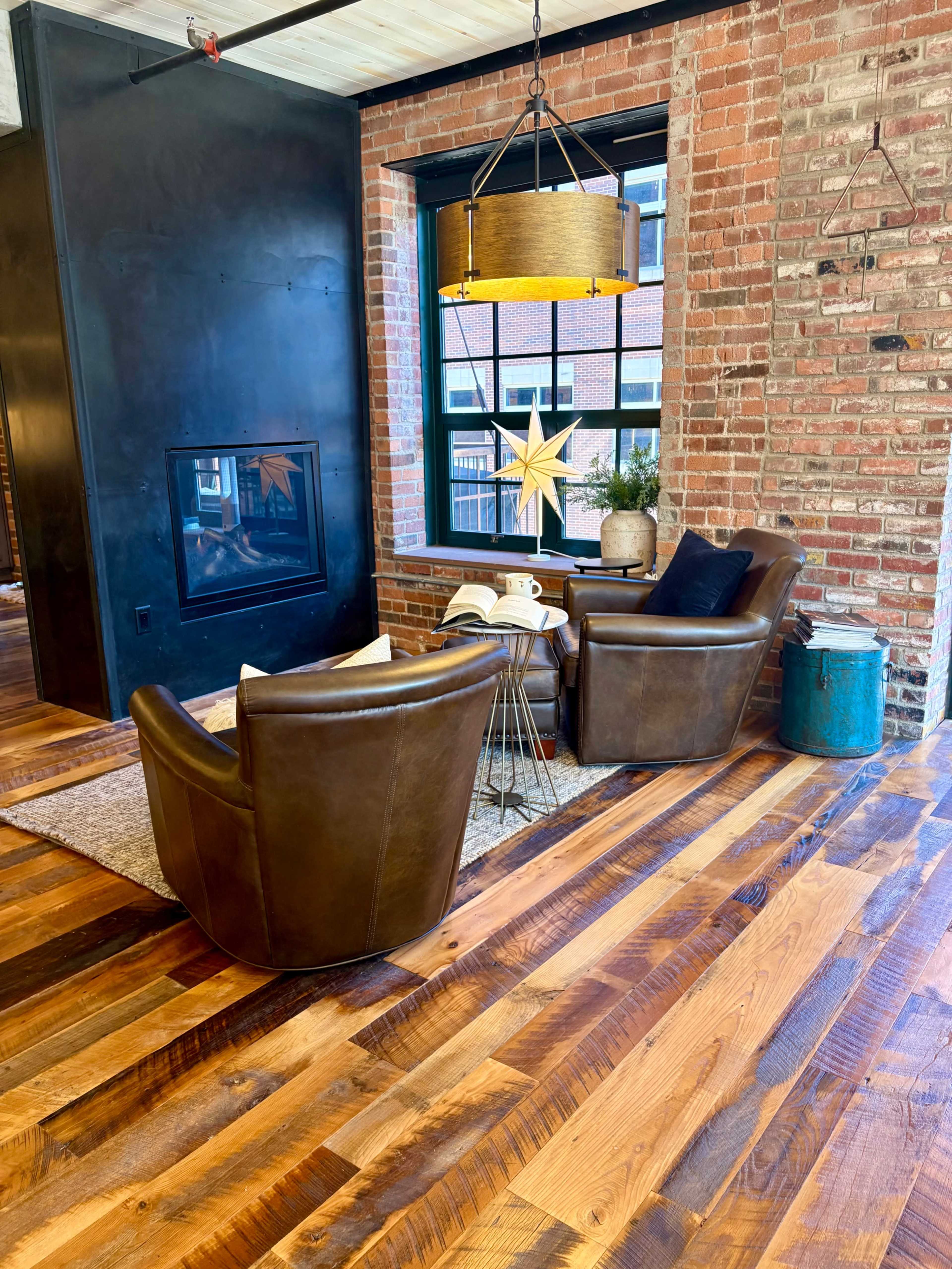The image shows a cozy seating area with two leather chairs and a small table, illuminated by natural light through a large window framed by exposed brick walls.