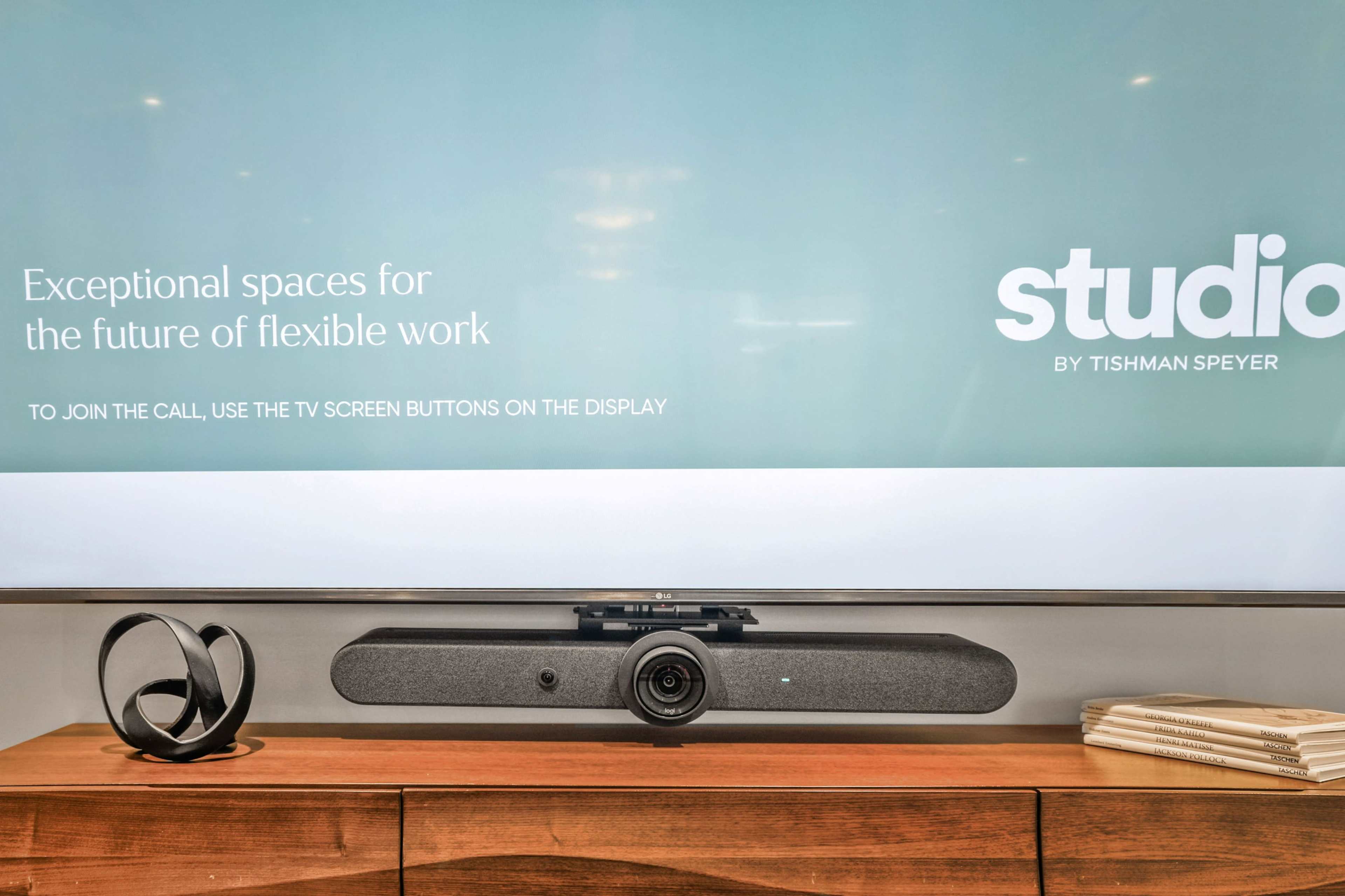 A television display shows a message about flexible work, with a video conferencing camera and speaker placed on a wooden surface below it.