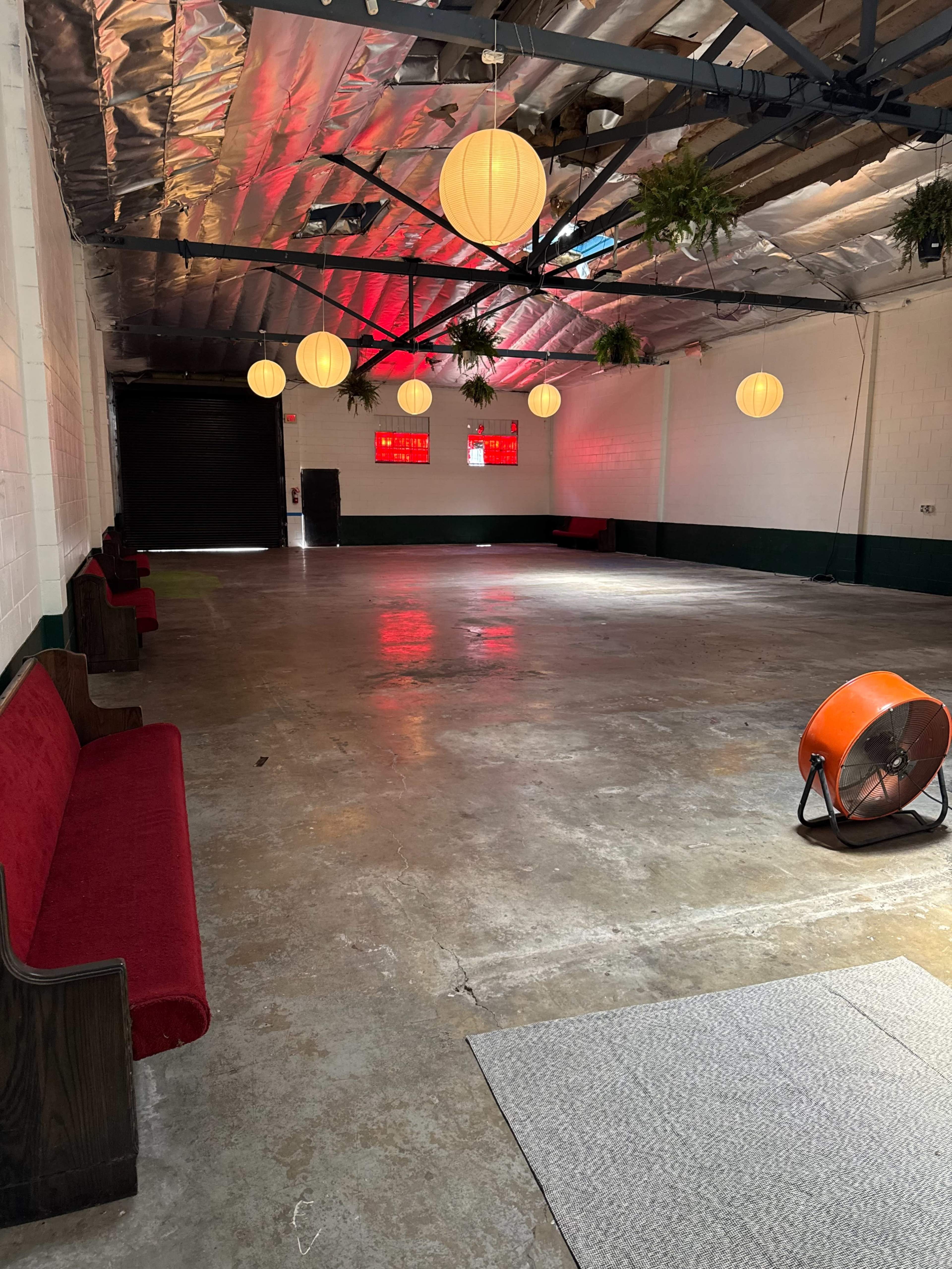 The image shows a spacious, empty indoor area with concrete flooring, red benches along the walls, and hanging lanterns providing soft lighting.