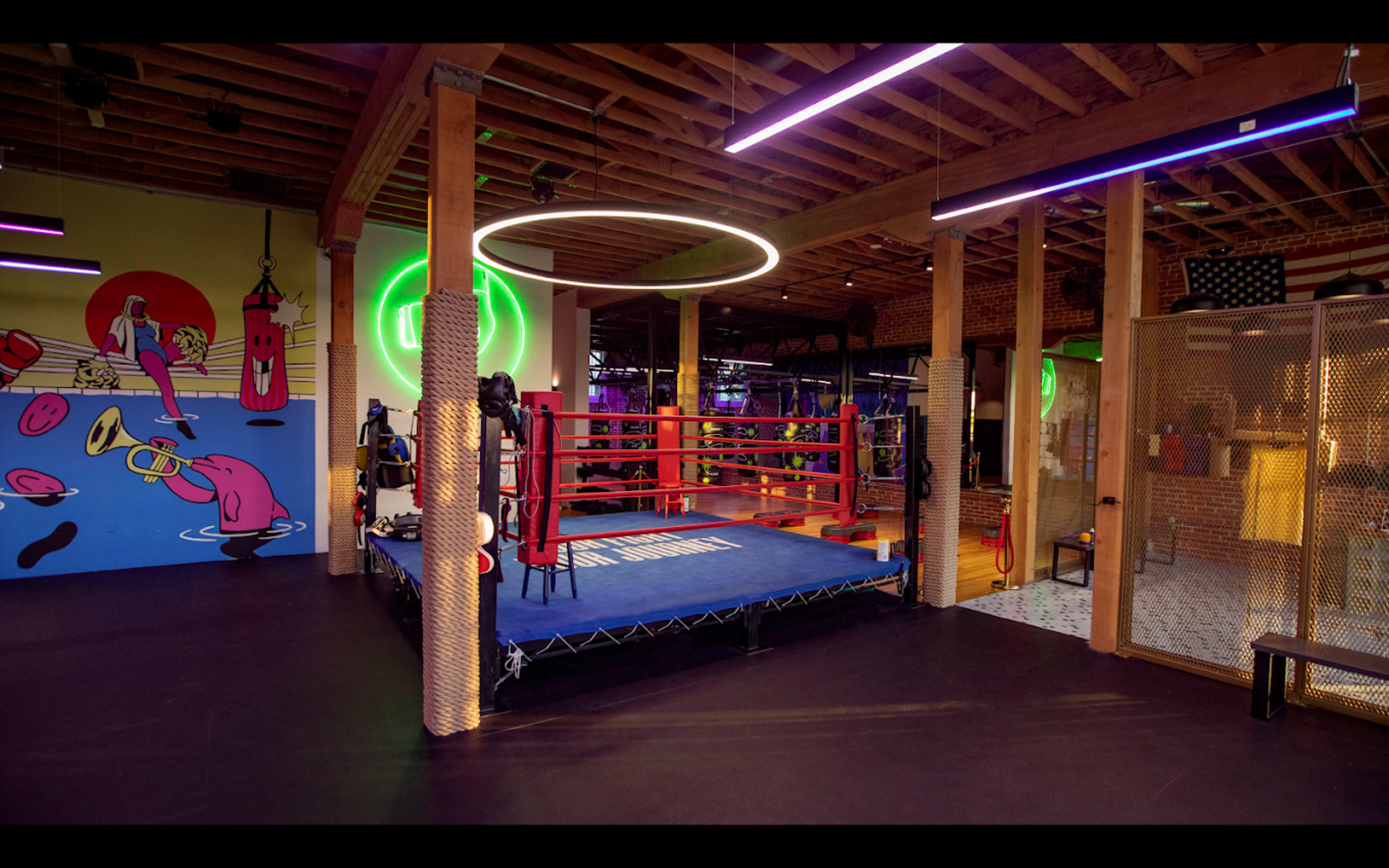 A modern boxing gym with a red boxing ring, colorful wall art, and various fitness equipment visible.