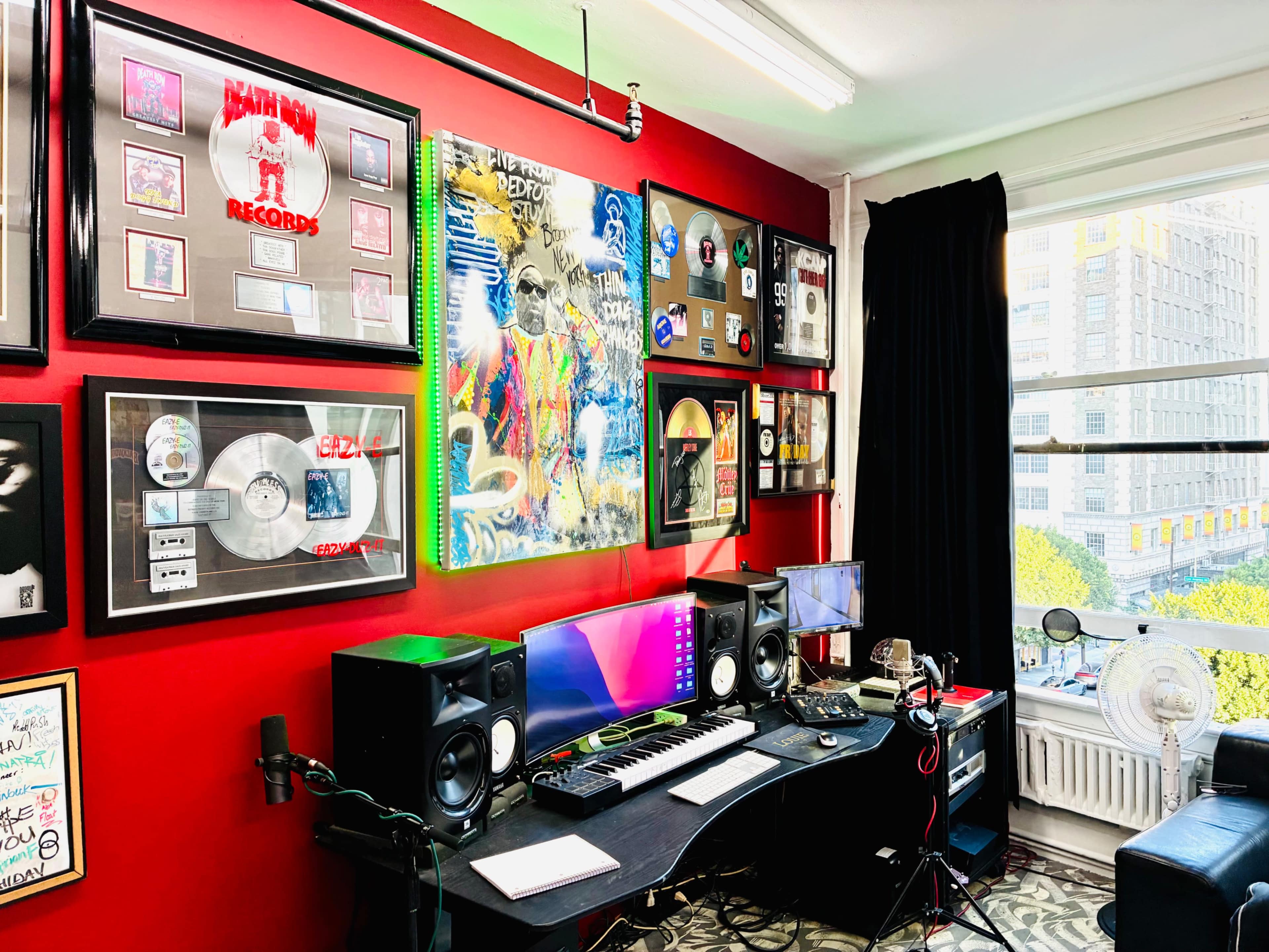 The image shows a music production studio featuring a black desk with computer monitors, music equipment, and a colorful wall adorned with framed records and art.