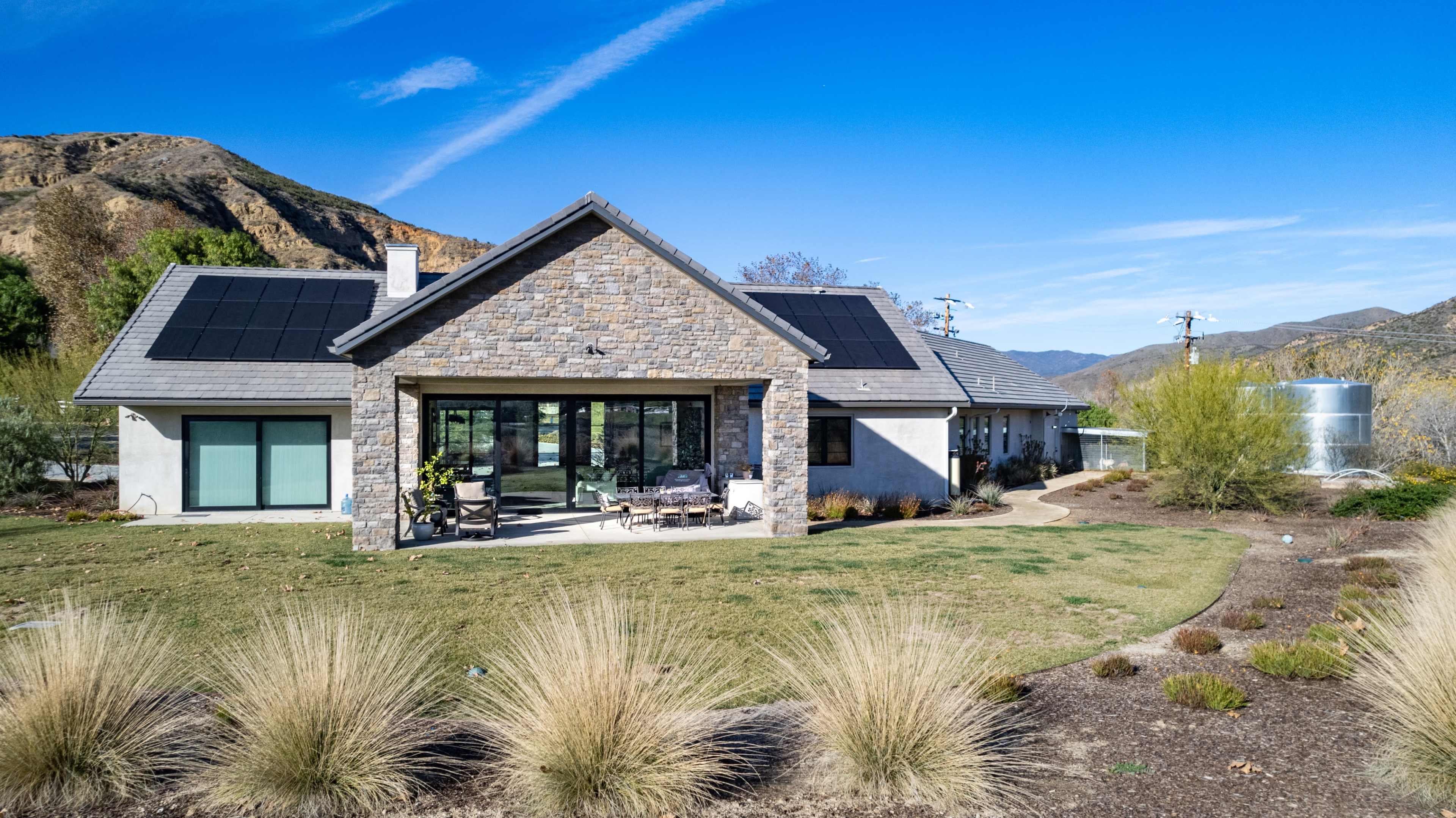 A modern house with solar panels on the roof is surrounded by landscaped gardens and mountains in the background.