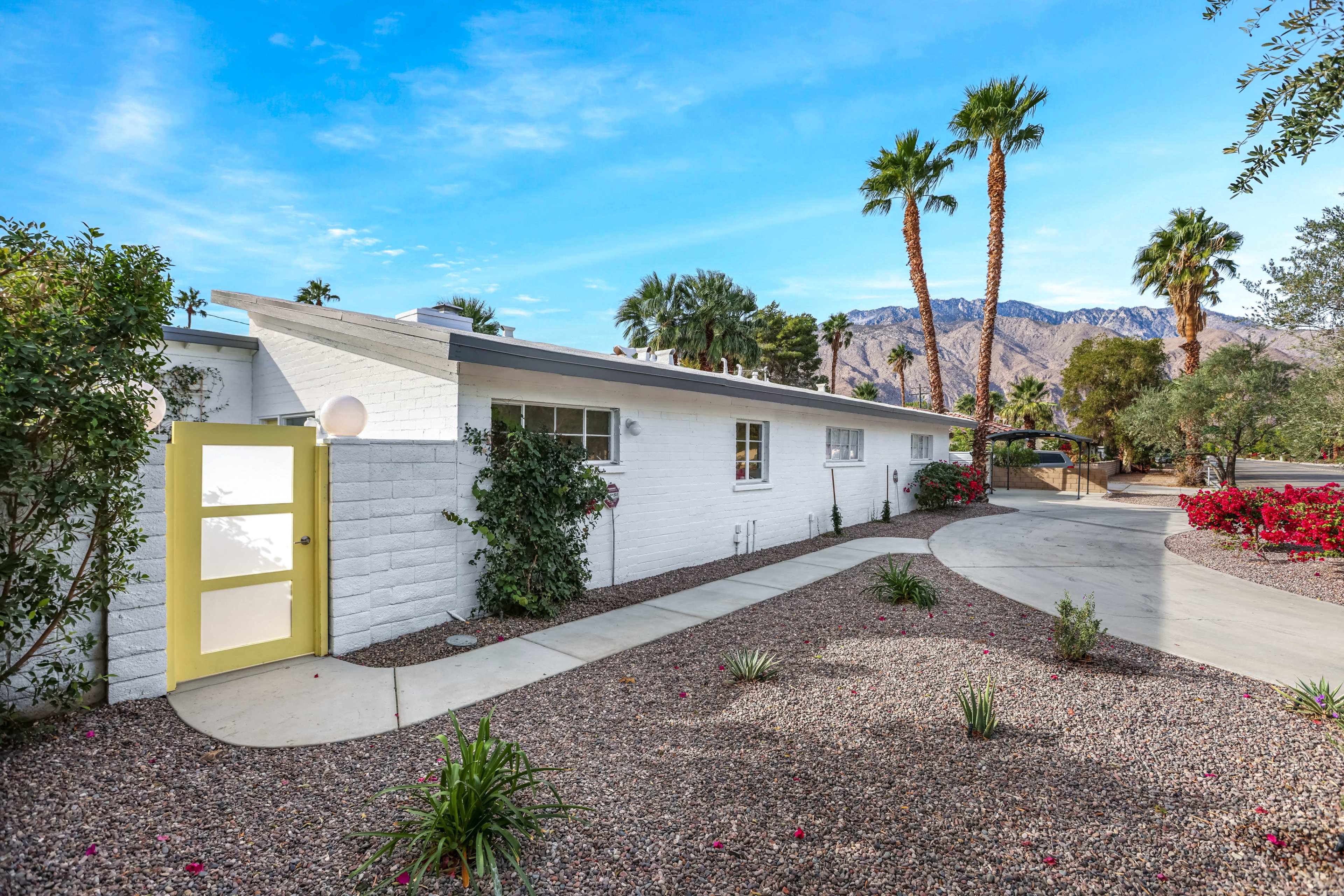A white house with a yellow door sits alongside a winding gravel path, surrounded by palm trees and lush landscaping in the foreground.
