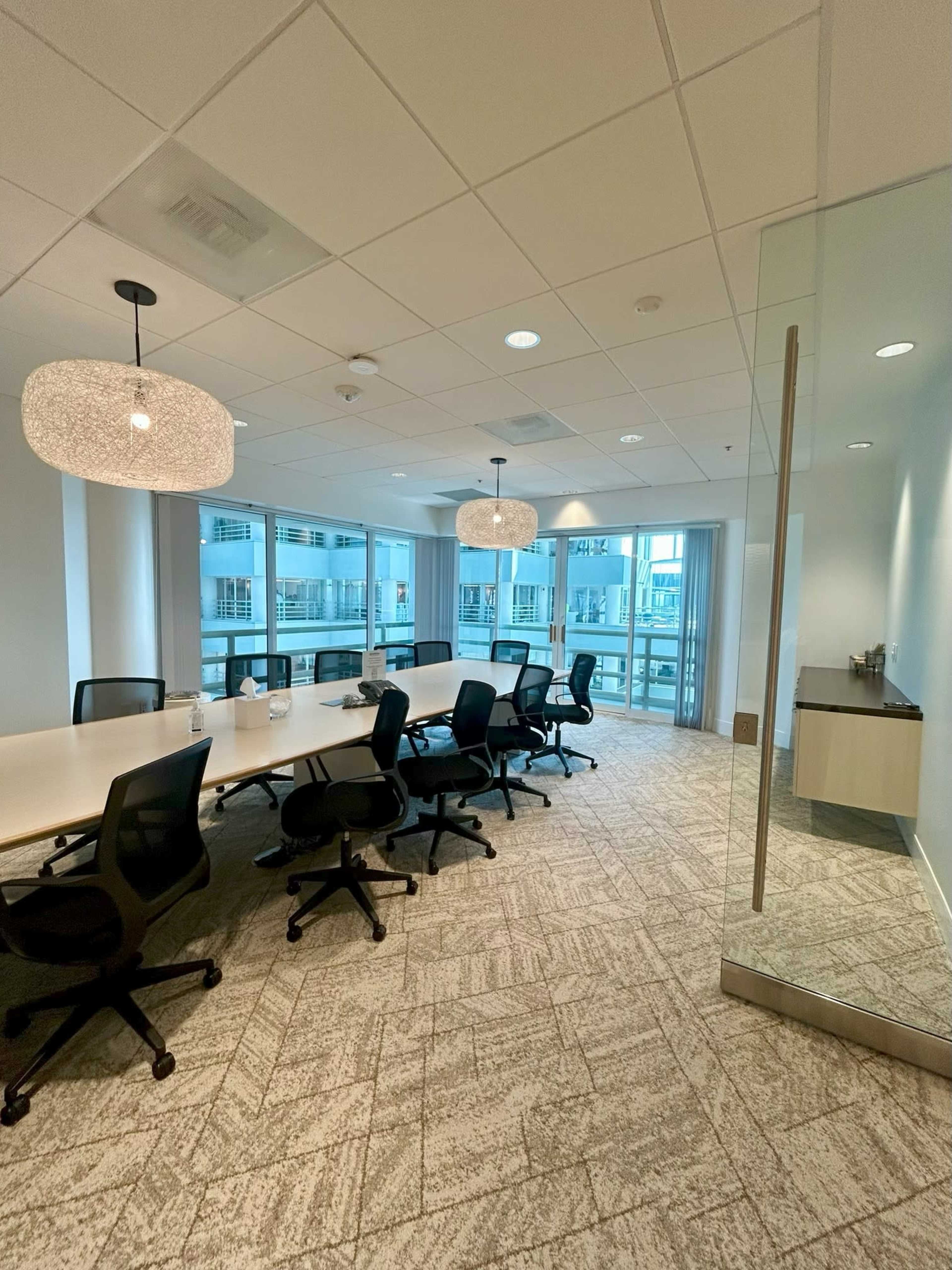 The image shows a modern conference room with a long table, black office chairs, and large windows overlooking a balcony area.