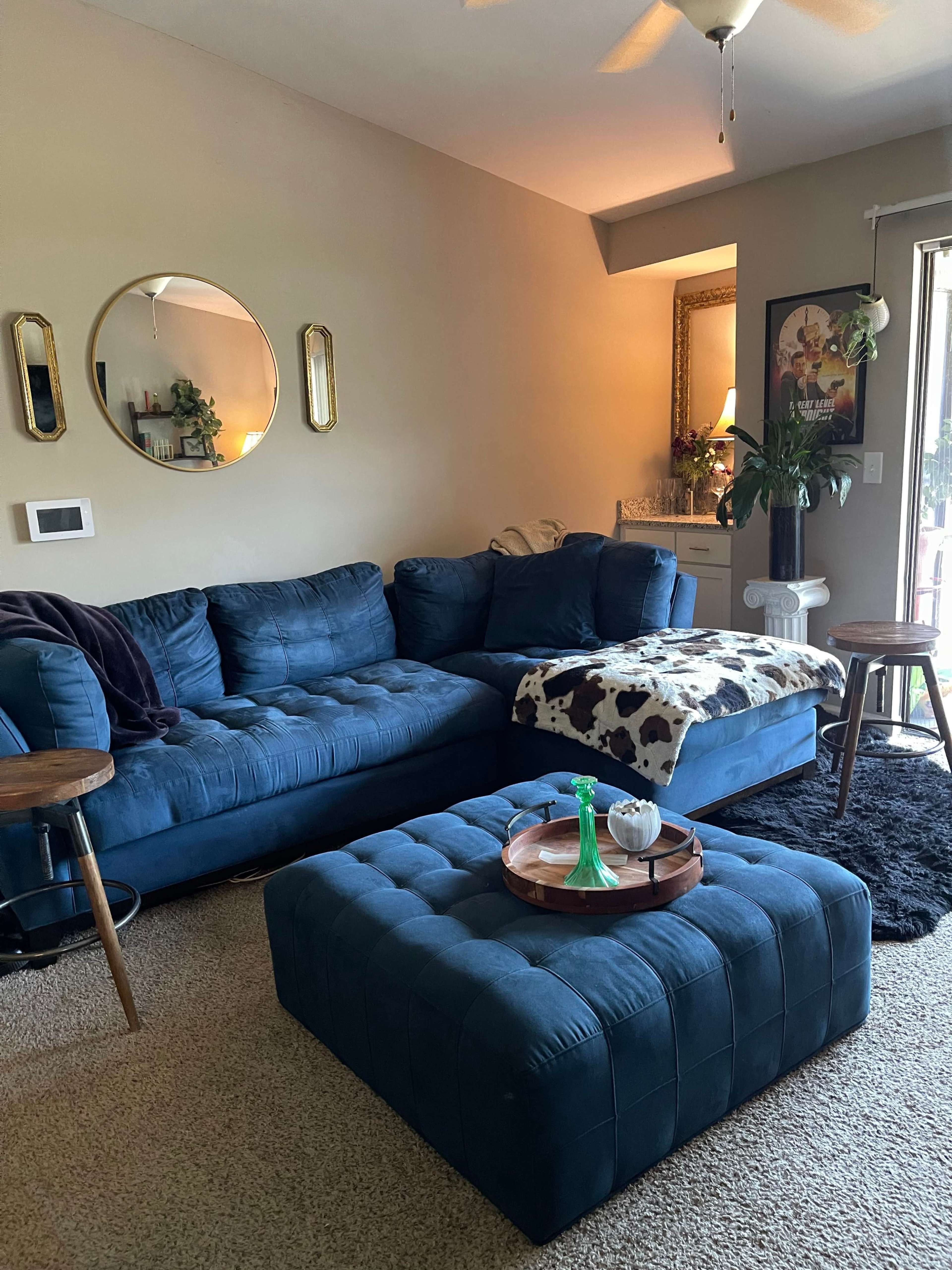 The image shows a cozy living room with a blue sectional sofa, a round mirror, a coffee table with decorative items, and a textured rug on the floor.