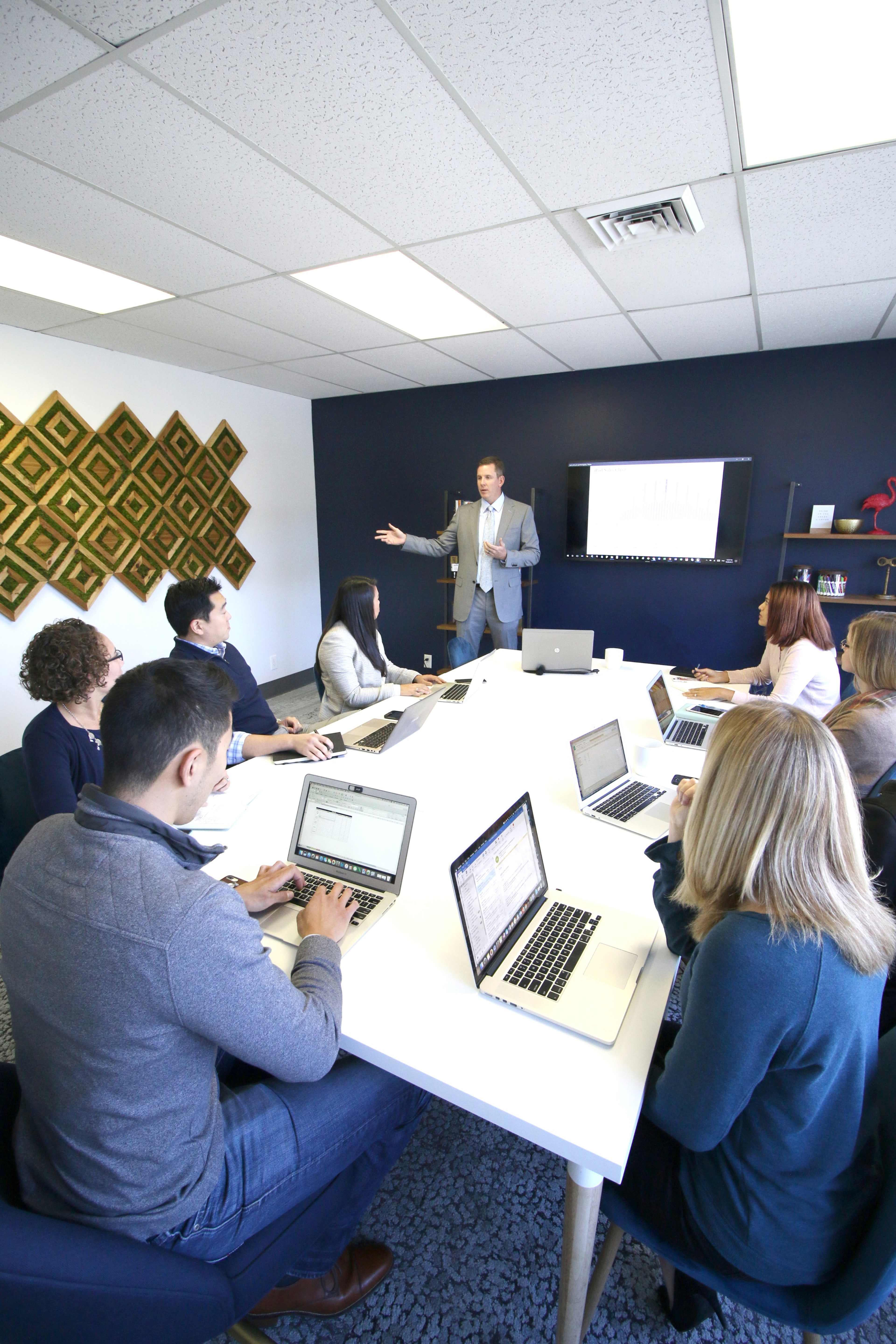 A group of eight professionals is engaged in a meeting around a large table, with one person presenting in front of a screen displaying data.