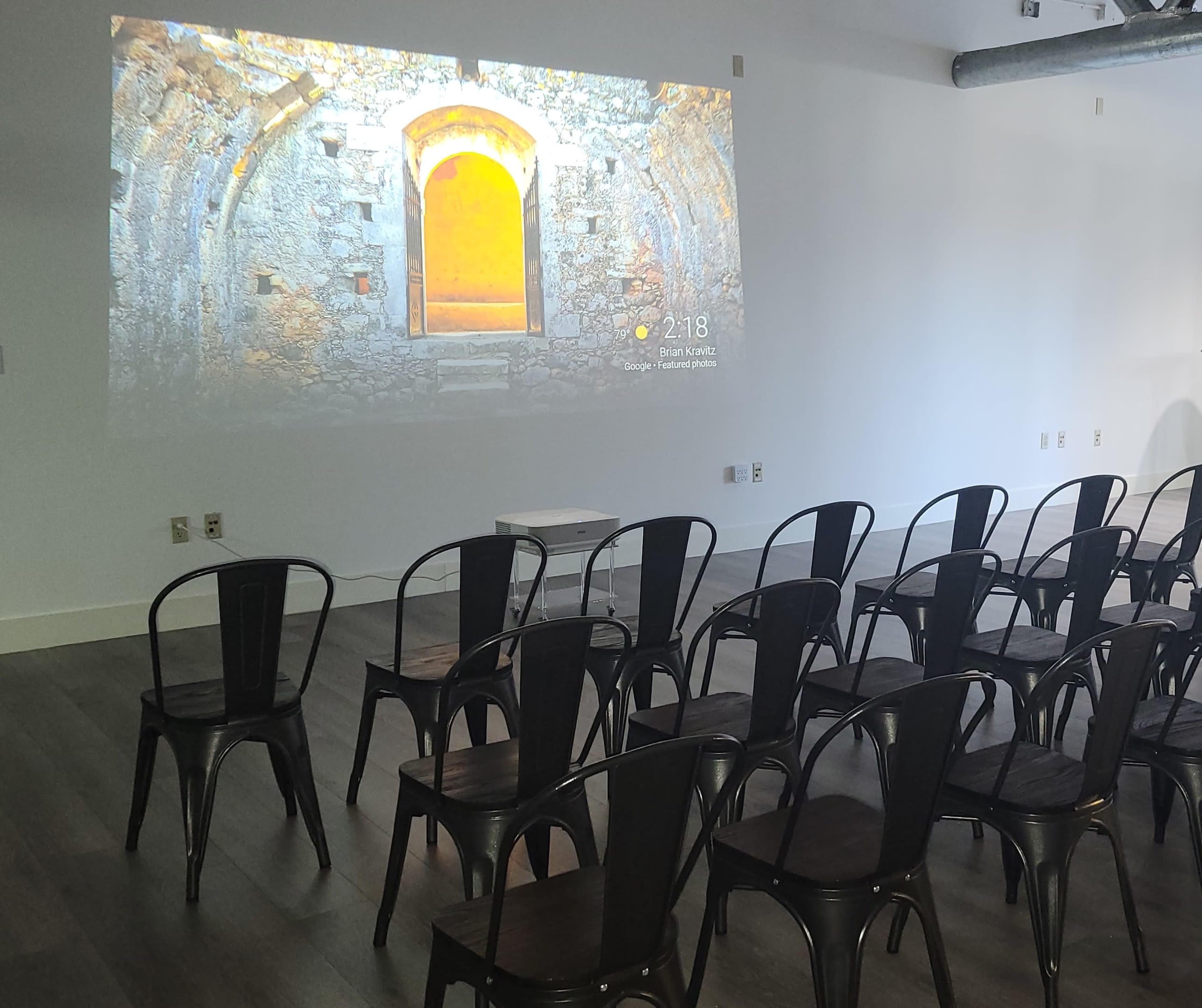 A room with rows of black metal chairs facing a projected image of a stone wall with a doorway.