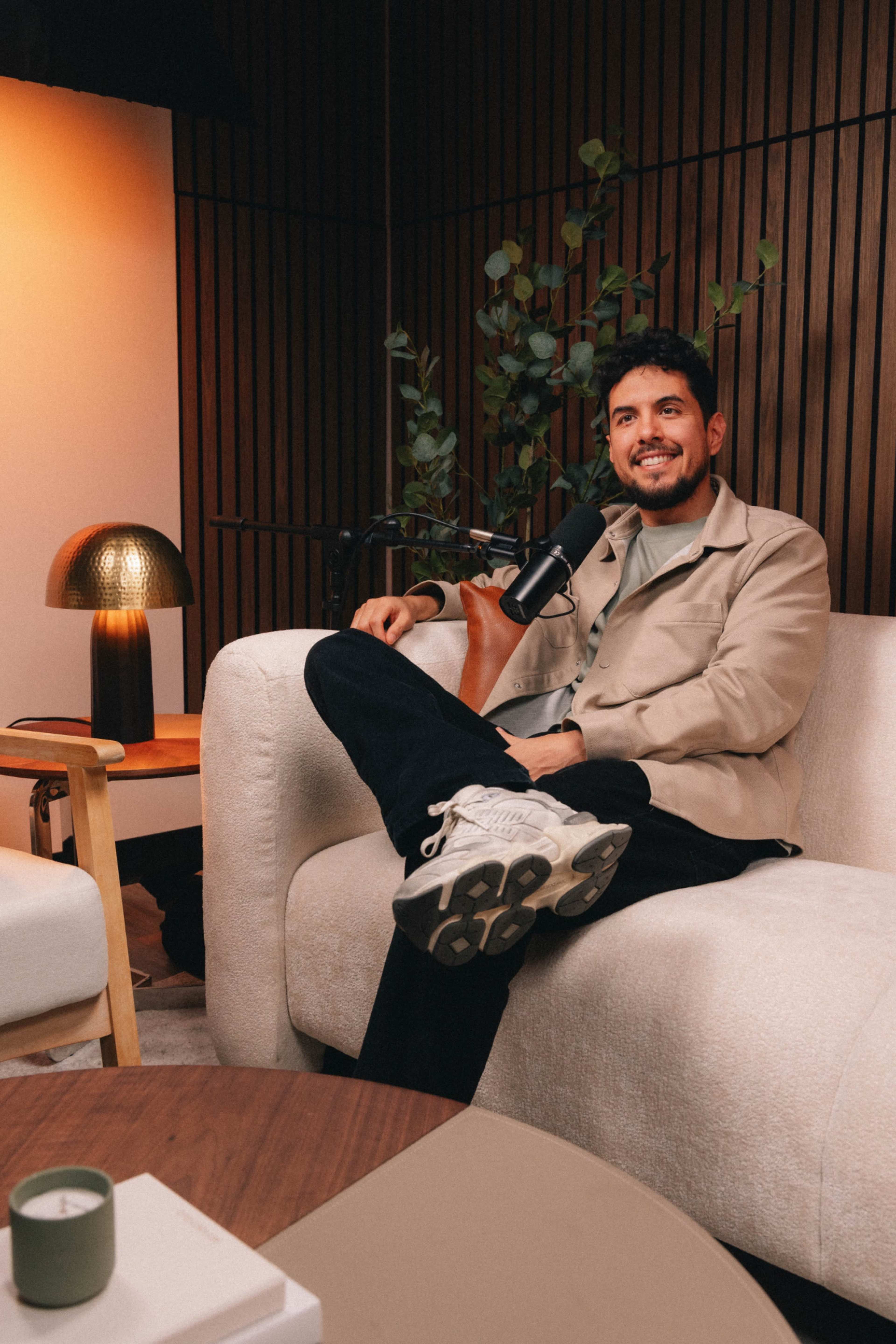 A man sits on a white sofa in a cozy interior, smiling while holding a microphone, with a plant and a lamp visible in the background.