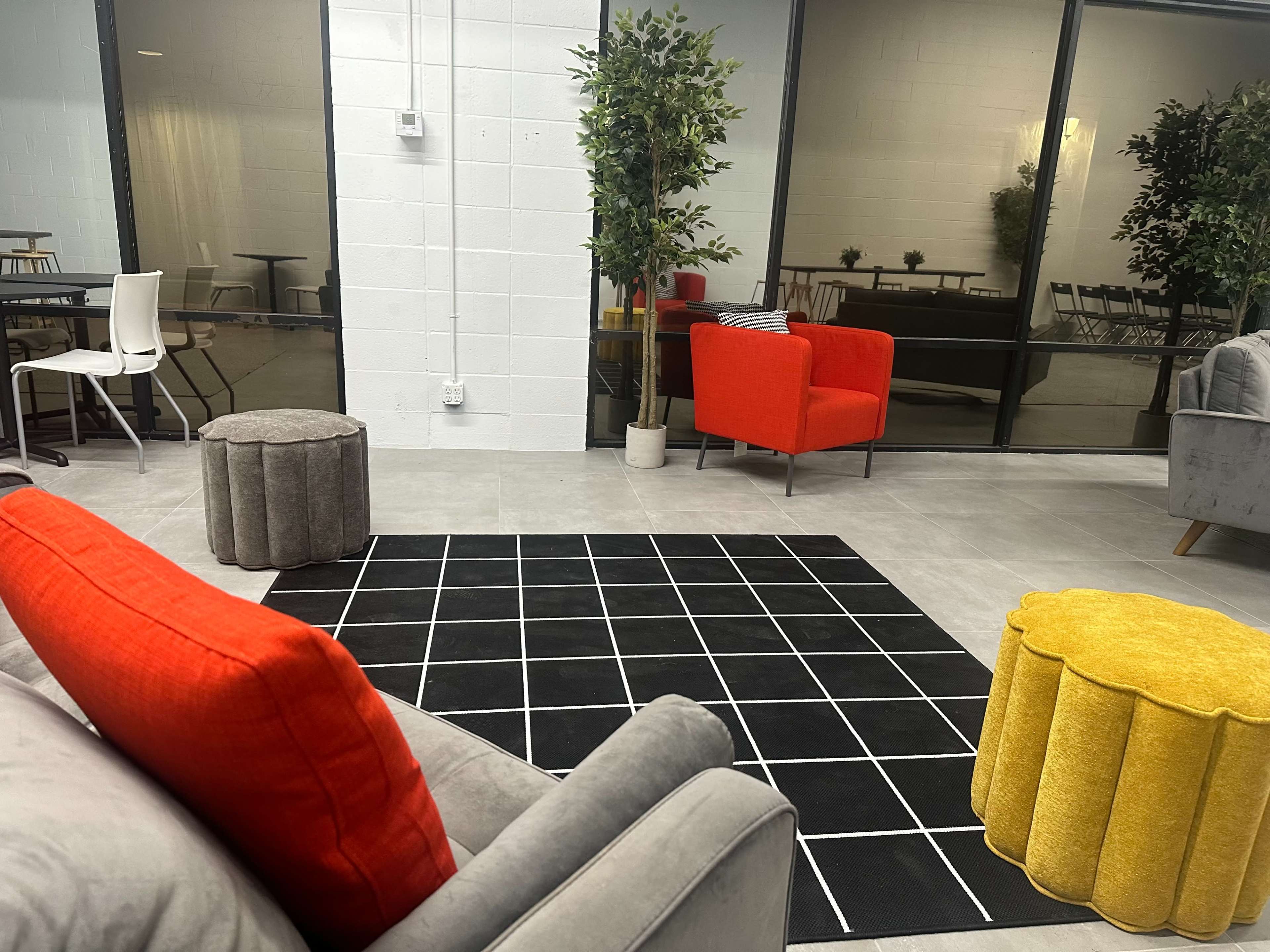 A modern lounge area with a gray sofa, an orange armchair, a yellow stool, and a patterned black and white rug, surrounded by potted plants and large glass windows.