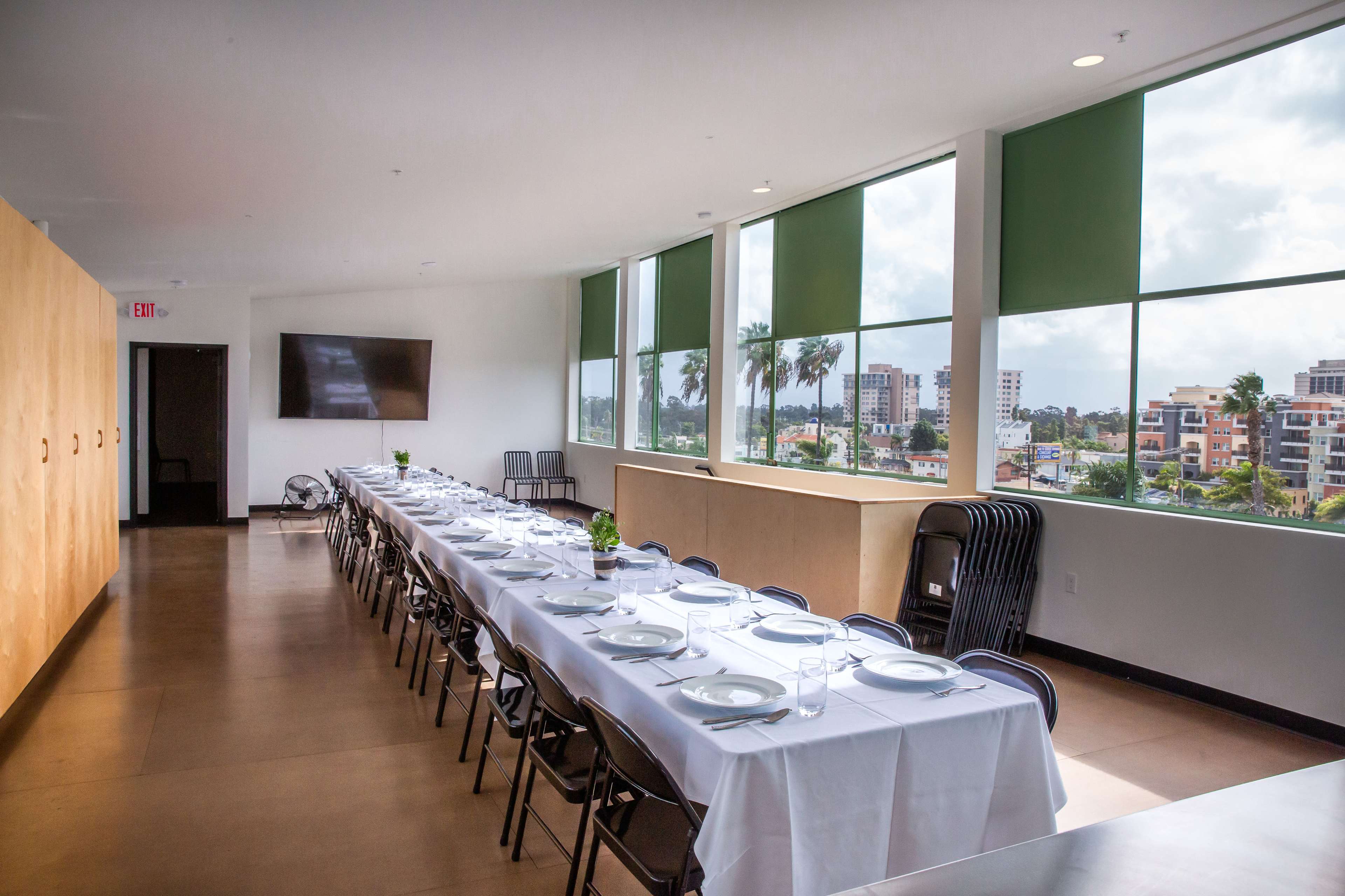 A long dining table is set with plates and glasses in a bright, modern room featuring large windows that overlook a cityscape.