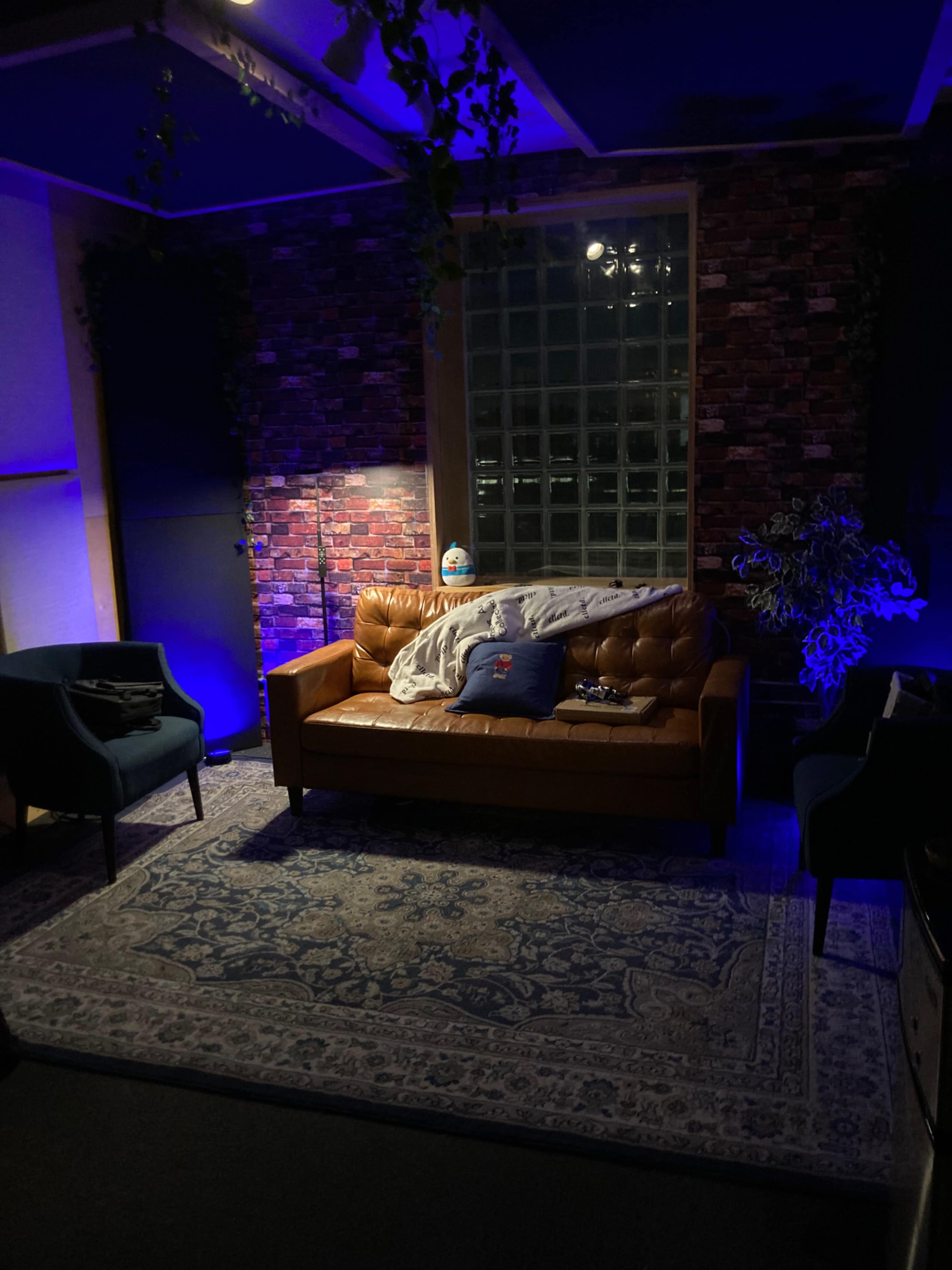 A dimly lit lounge area featuring a brown leather sofa, two blue chairs, a decorative rug, and potted plants against a brick-patterned wall.