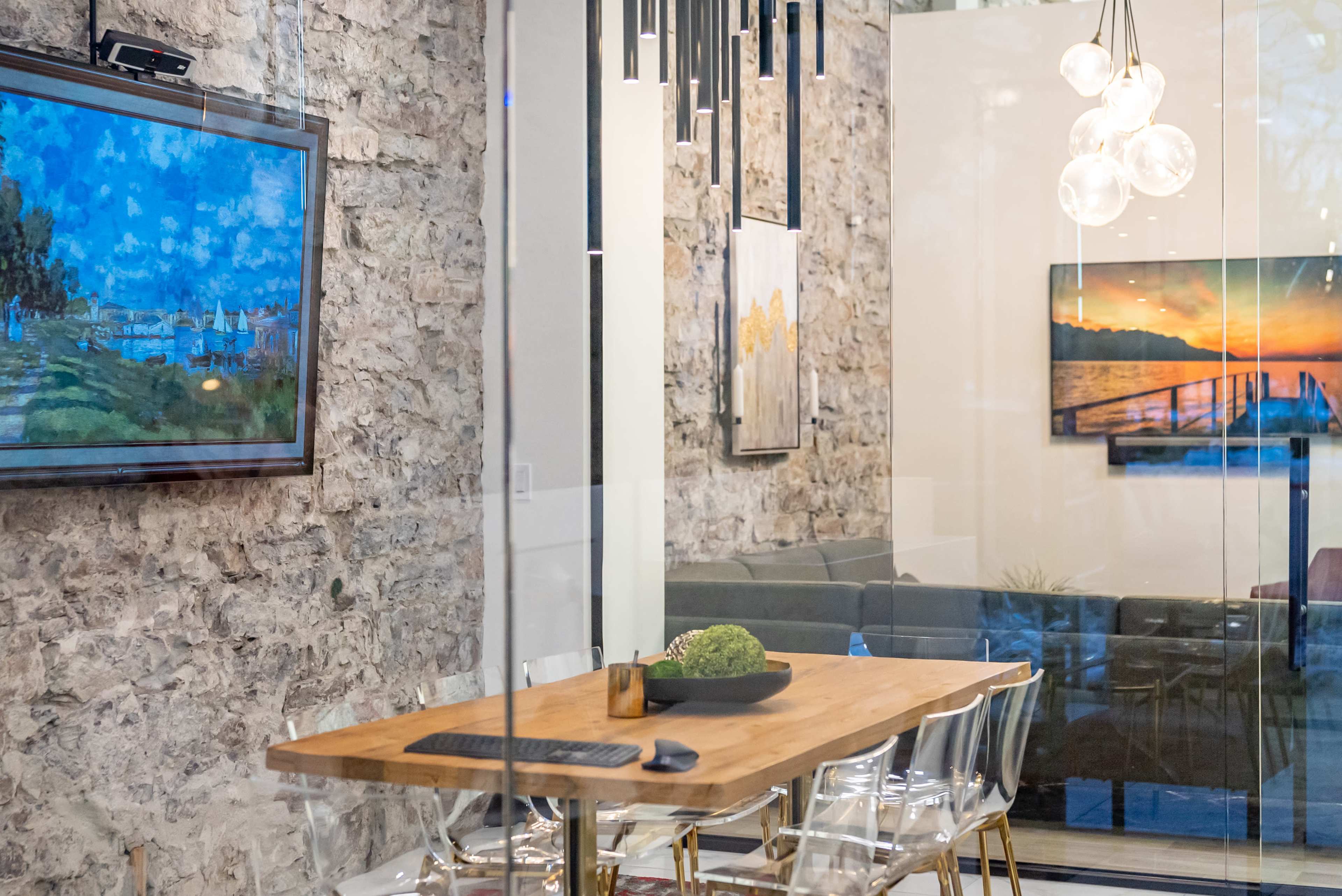 A modern meeting room features a wooden table surrounded by clear chairs, stone walls, and framed artwork, with a television mounted on one wall.
