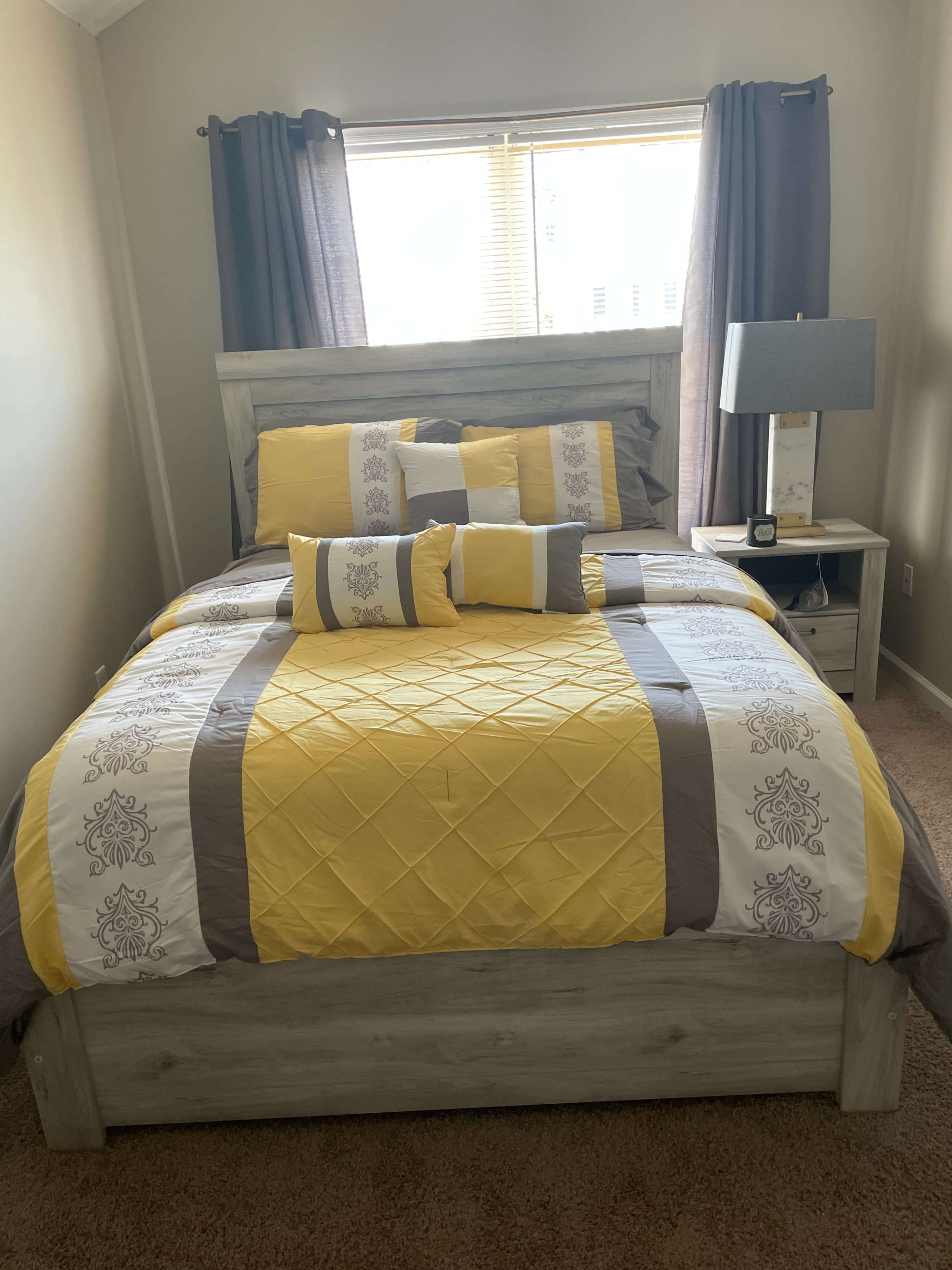 The image shows a neatly made bed with a yellow and gray patterned comforter and decorative pillows, positioned in a well-lit bedroom with curtains drawn partially open.