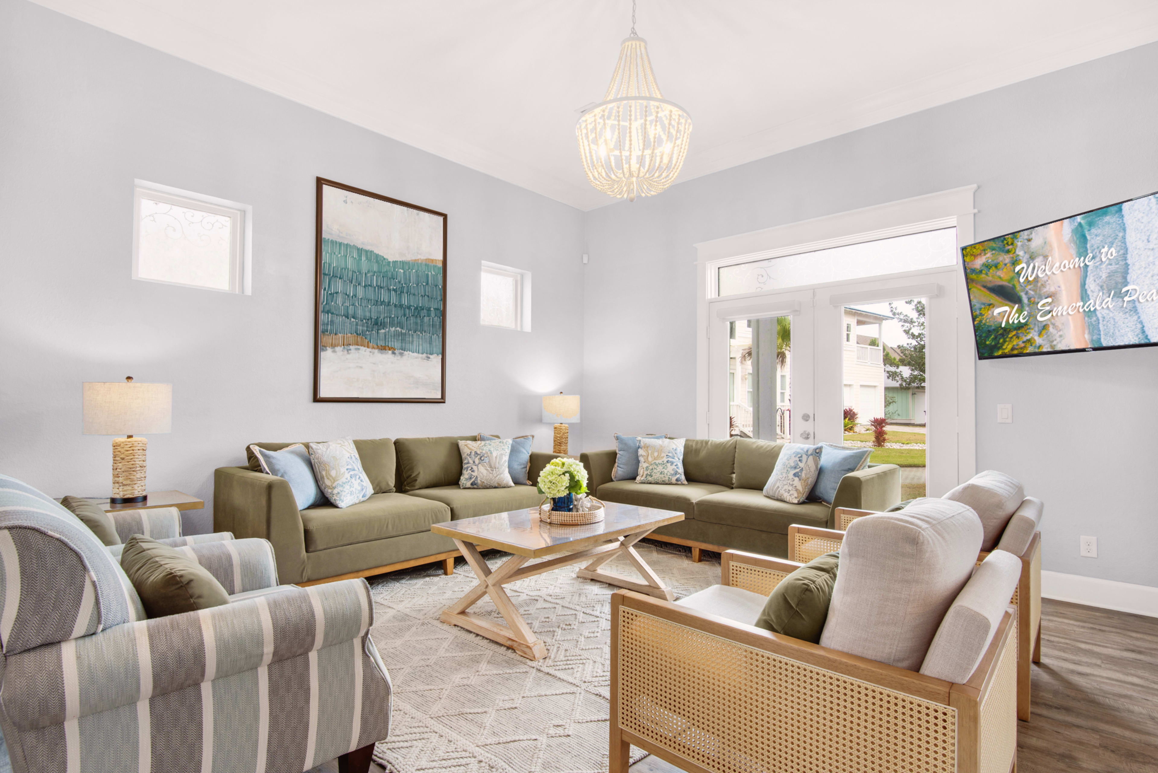 A spacious living room features two green sofas, light chairs, a wooden coffee table, and a large window with a decorative light fixture above.