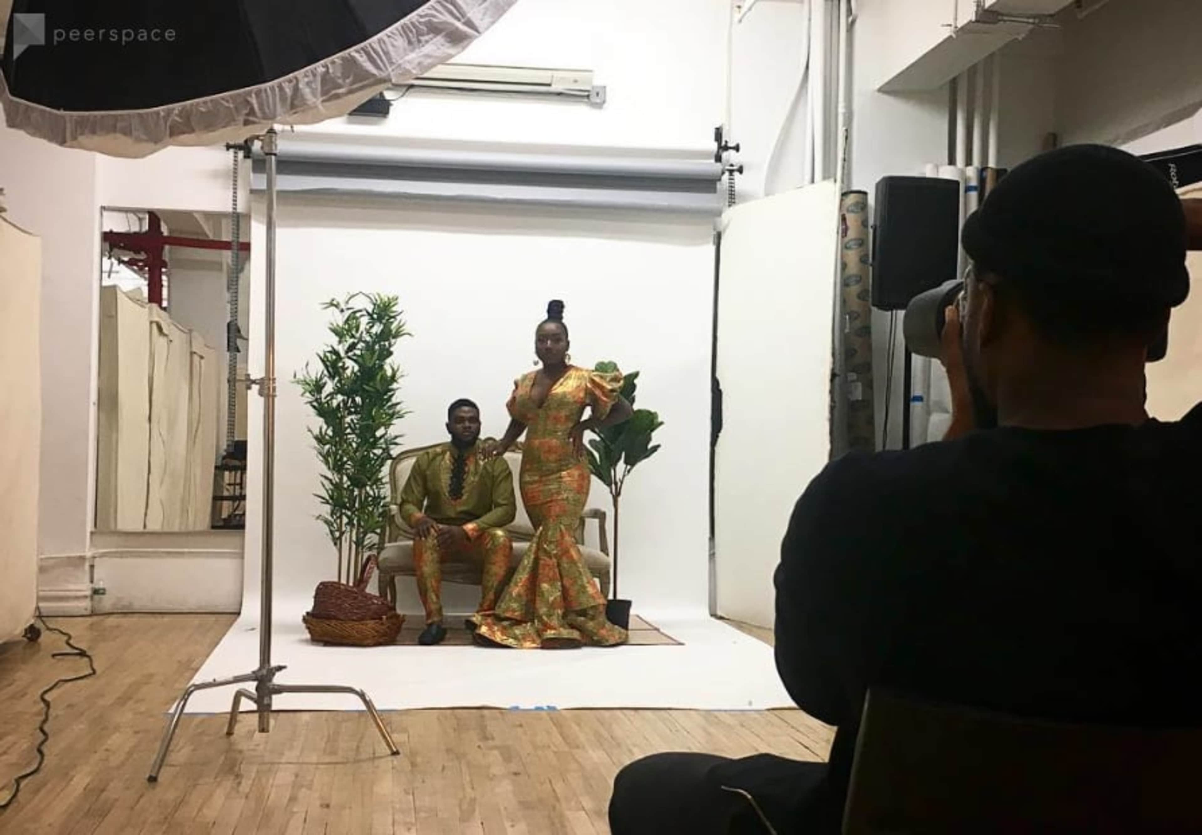 A photographer captures a couple posing on a couch in a studio setting with plants and a backdrop.
