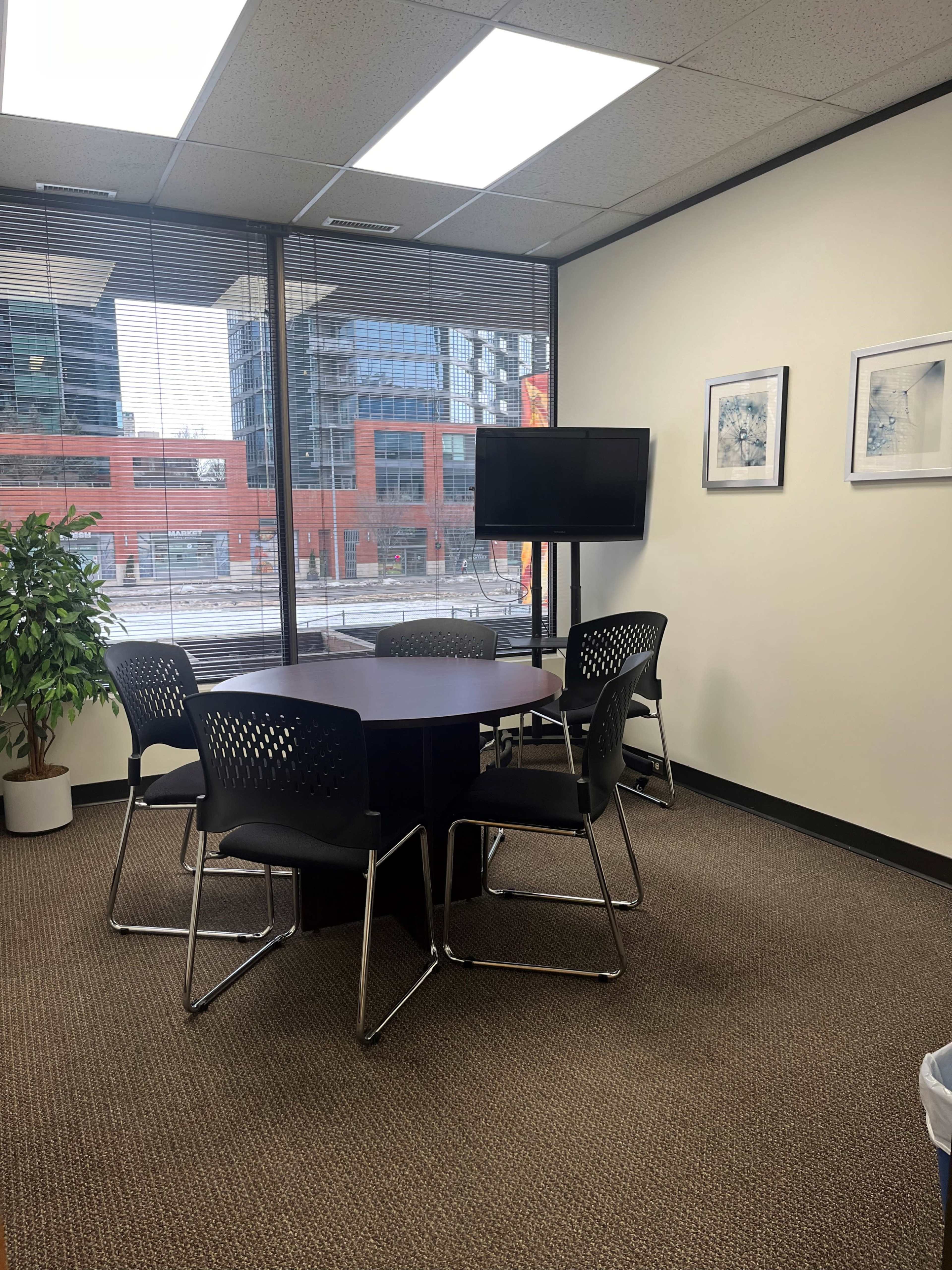 The image shows a small conference room with a round table and four black chairs, featuring large windows overlooking a cityscape and a television mounted on the wall.