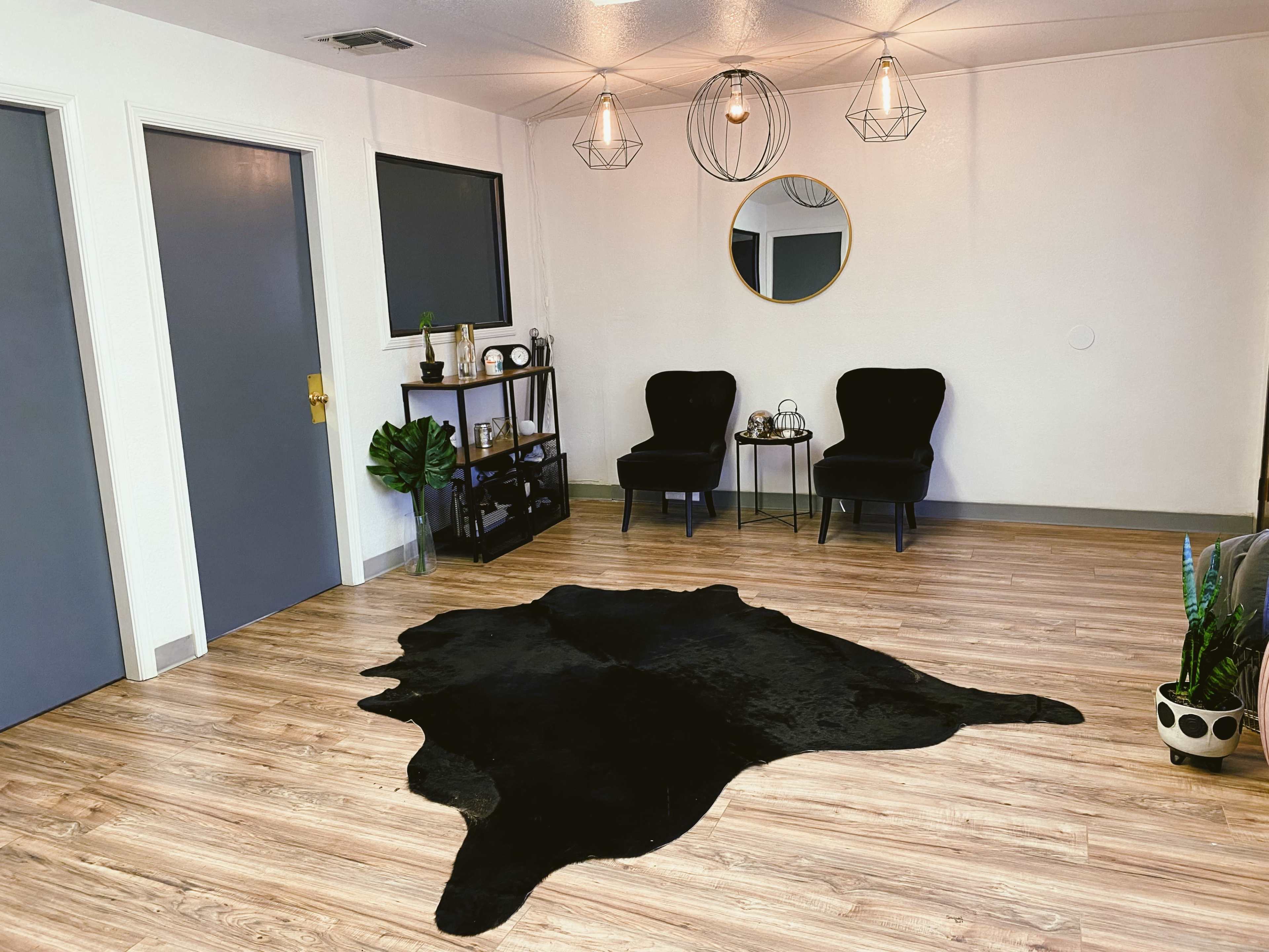 A minimalist room with two black chairs, a round mirror, and a black rug on wooden flooring, accompanied by a small shelving unit and potted plants.