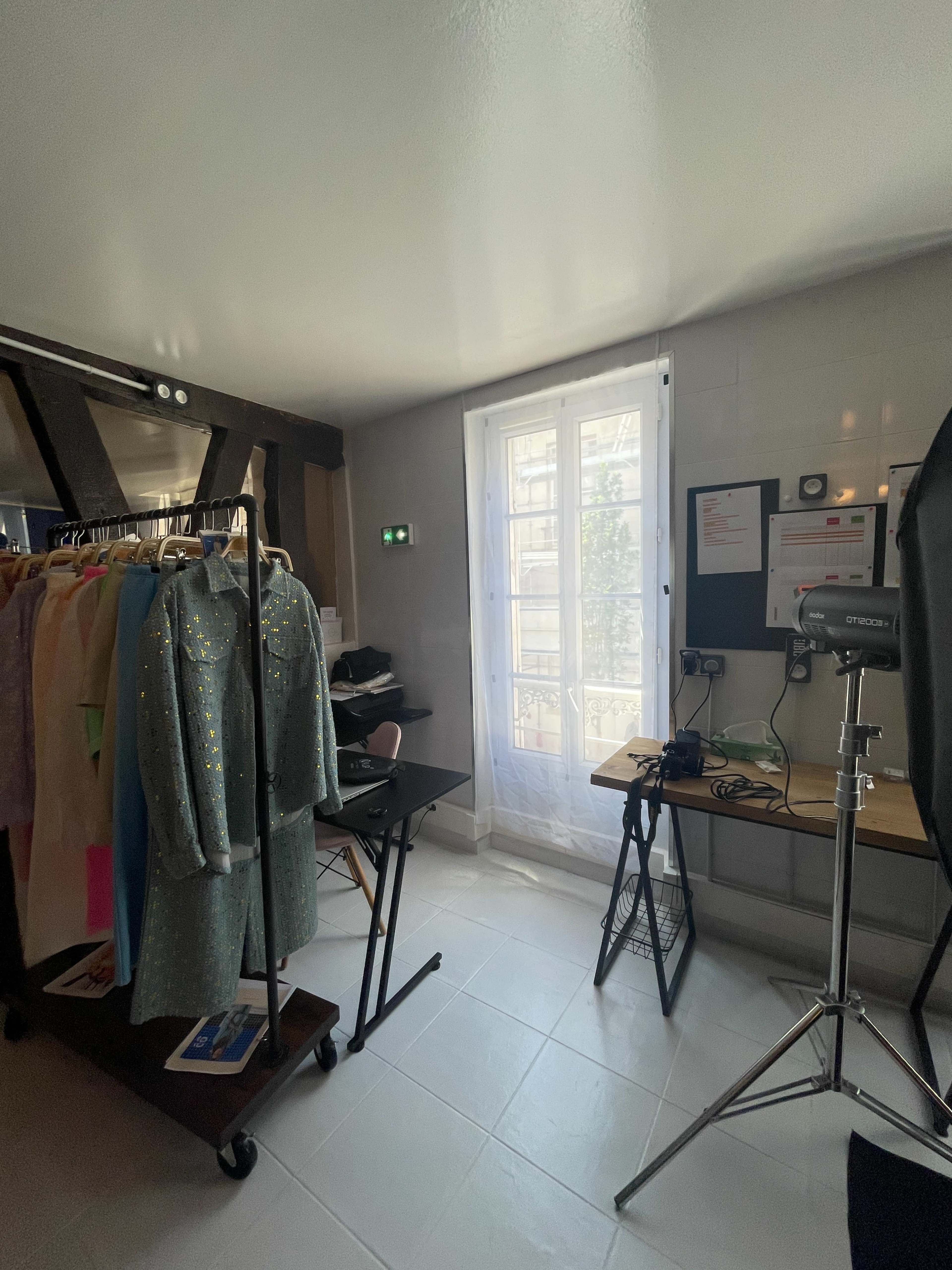 The image shows a small, well-lit room featuring a rack of colorful clothing, a desk with a computer setup, and a window illuminated by natural light.