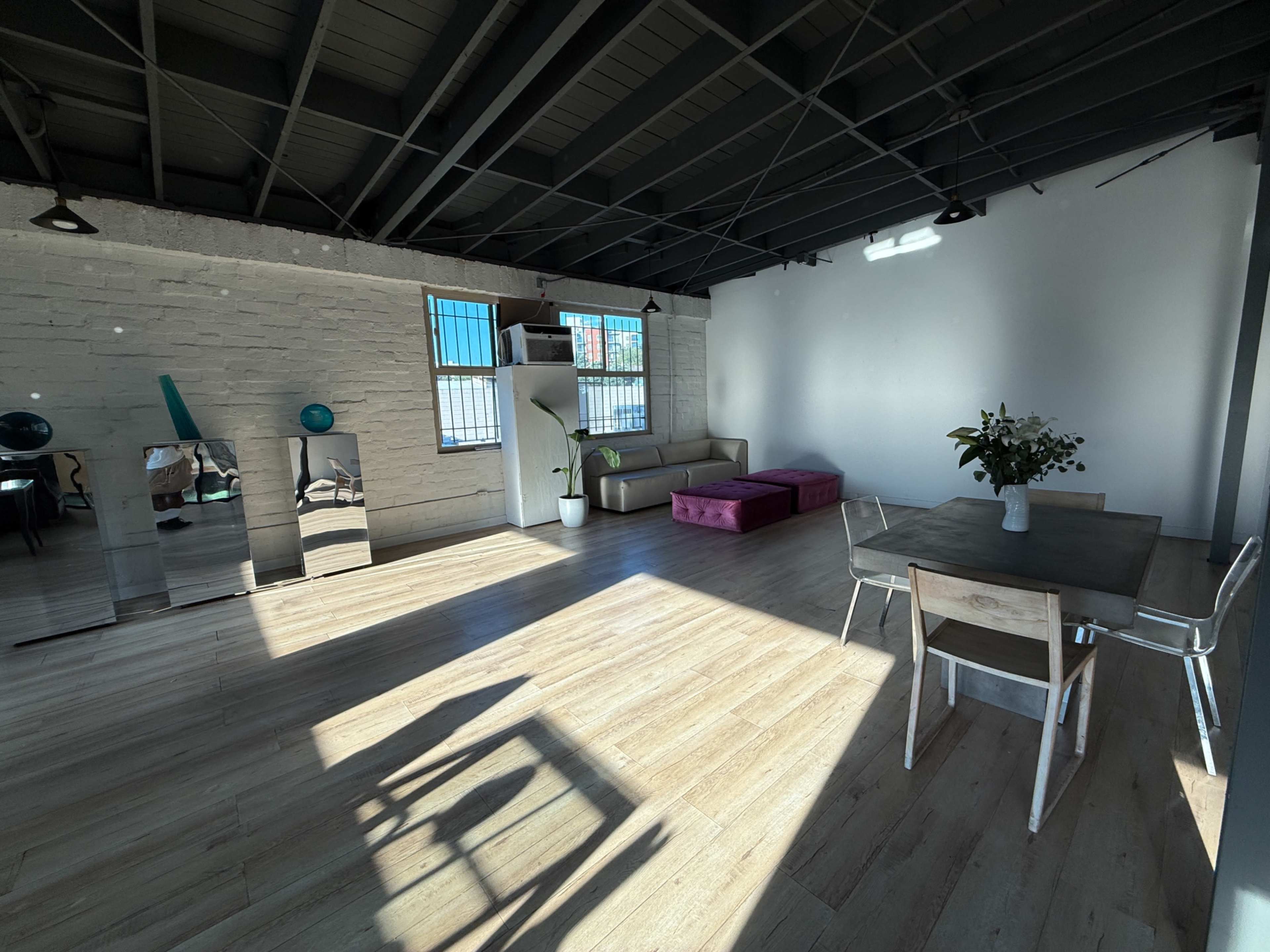 The room features a modern, open living space with a sofa, a purple ottoman, a dining table, and large windows allowing natural light to fill the area.