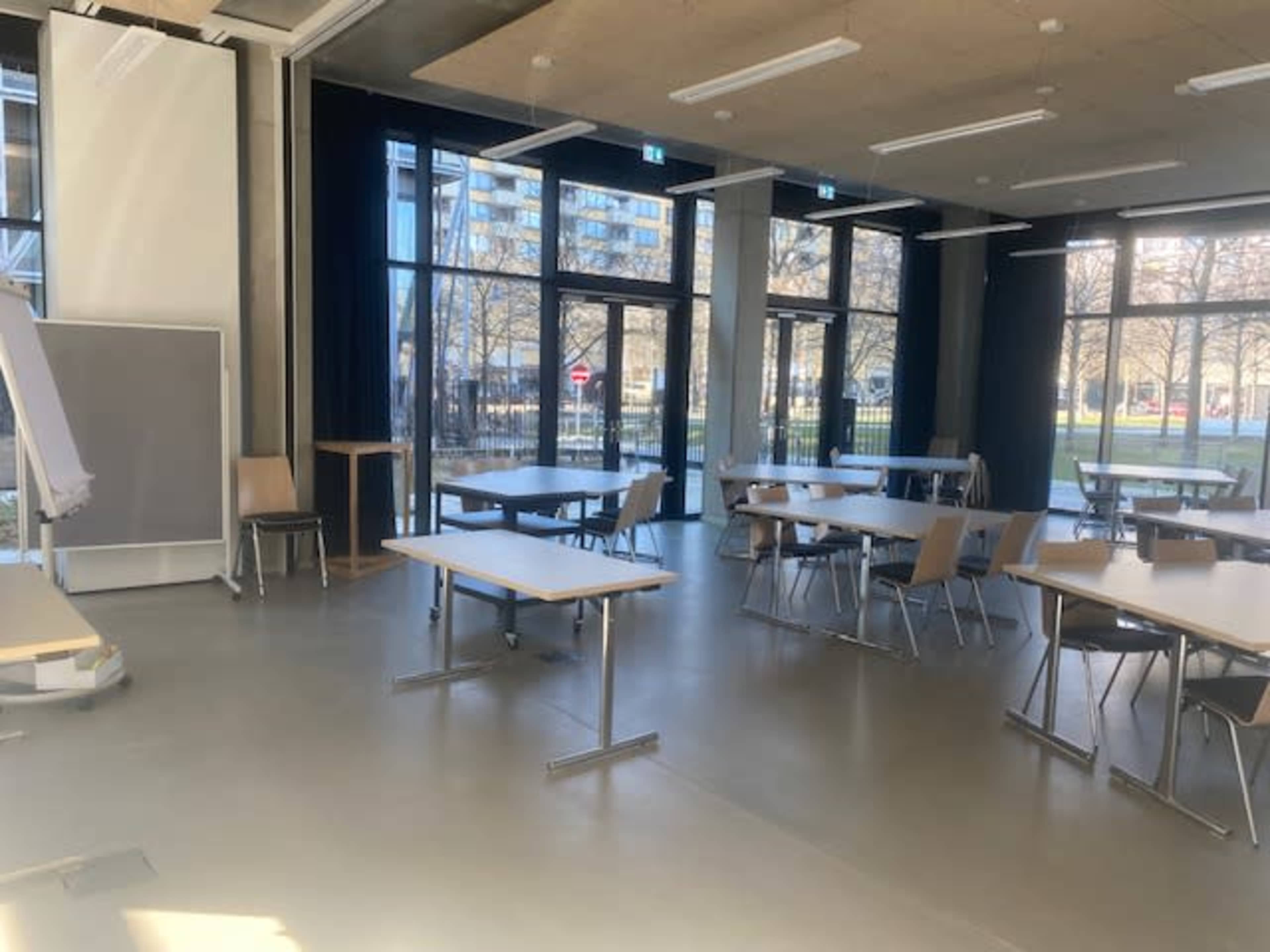 The image shows an empty classroom with tables and chairs arranged in a spacious layout, featuring large windows that overlook an outdoor area.