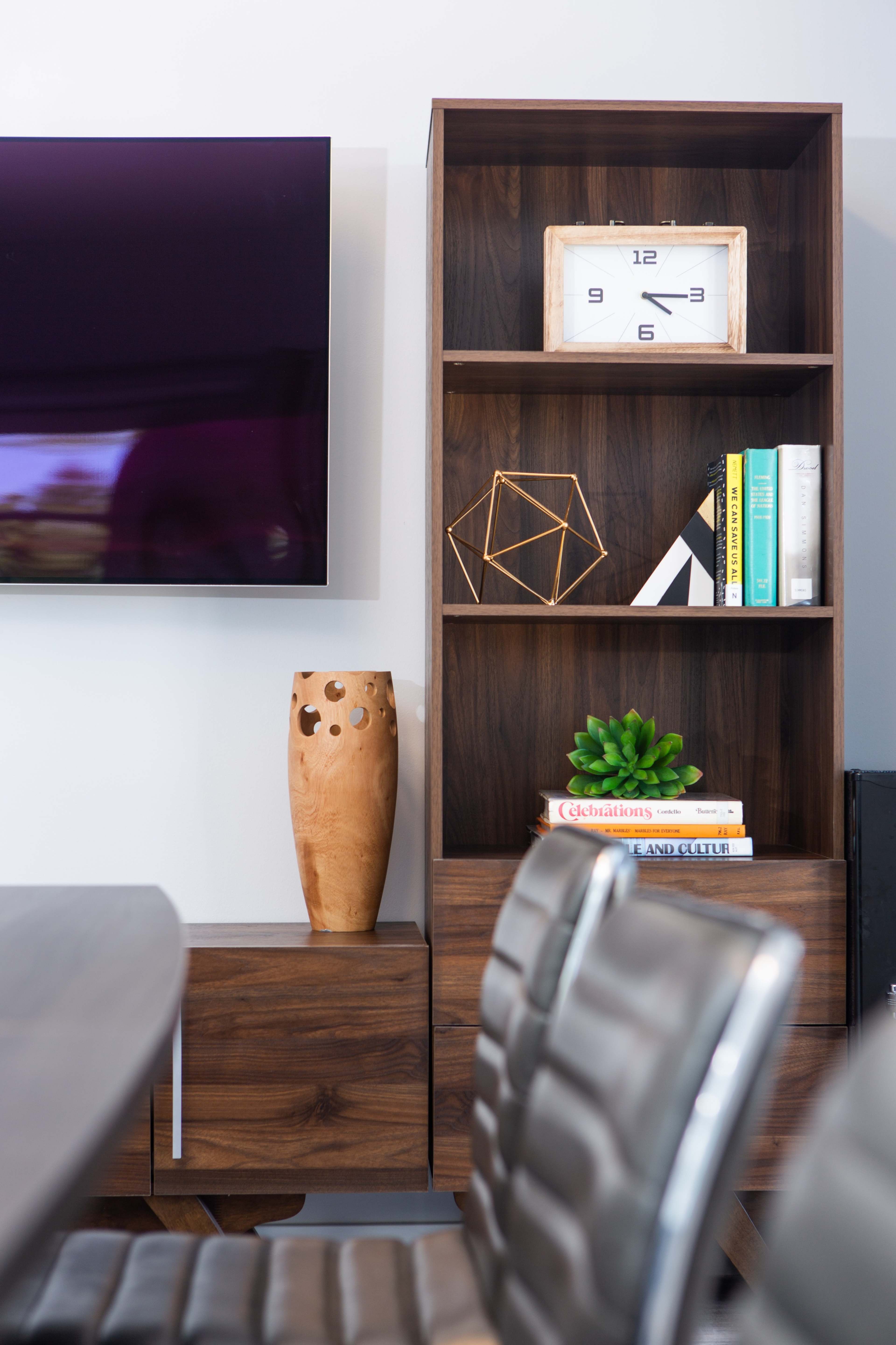 A wooden shelf displays a clock, a decorative geometric shape, books, a potted plant, and a carved vase beside a wall-mounted television.
