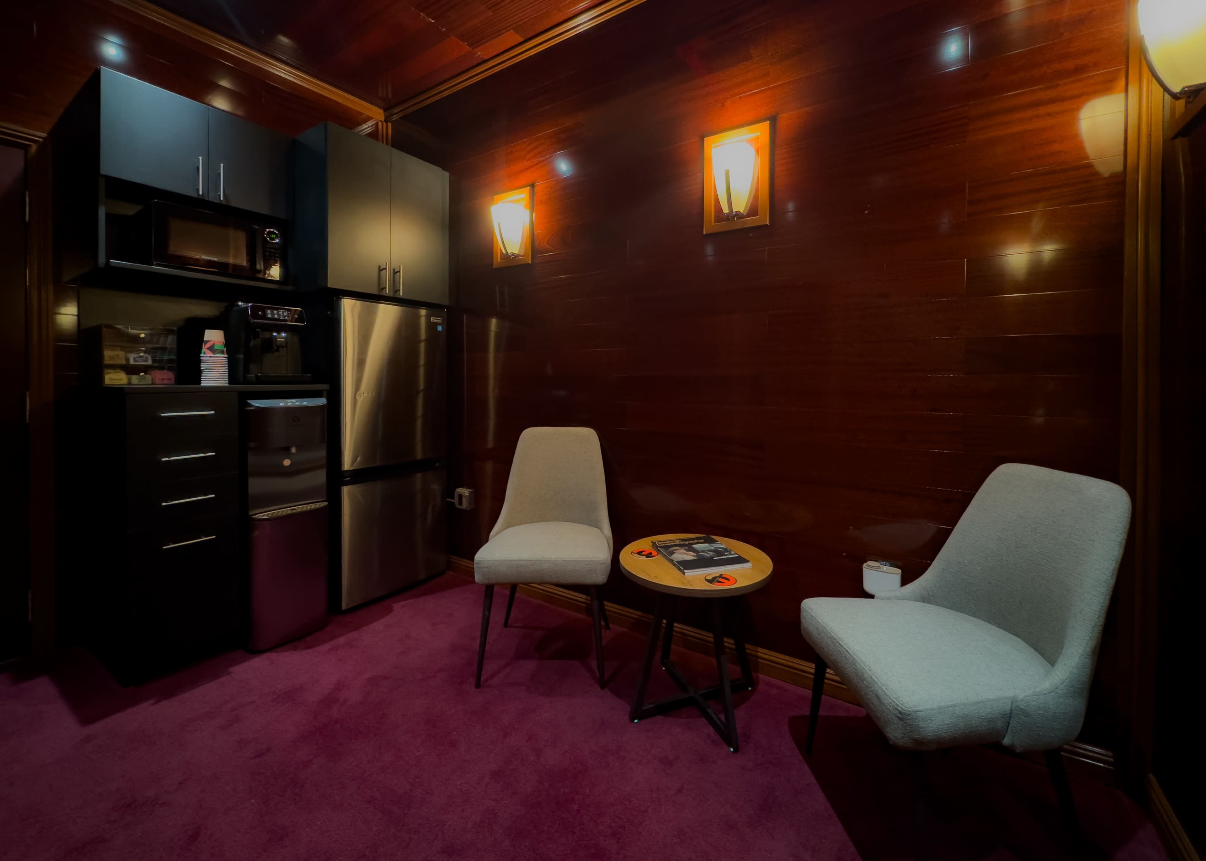 A small, cozy room with a dark wooden wall, a kitchenette featuring a refrigerator and microwave, and two gray chairs beside a round table on a purple carpet.