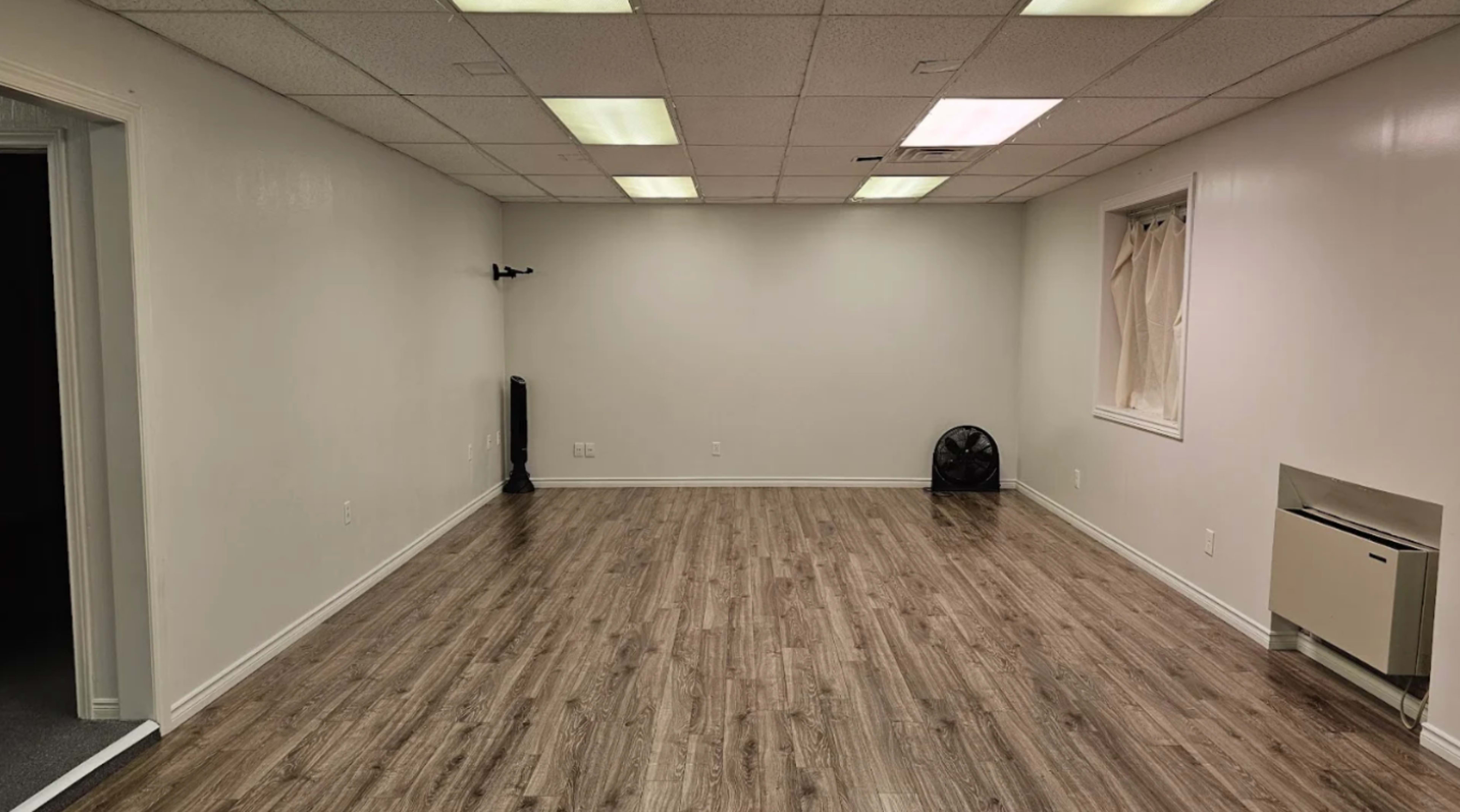 The image shows a sparsely furnished room with wooden flooring, white walls, fluorescent ceiling lights, and a small window with a curtain.