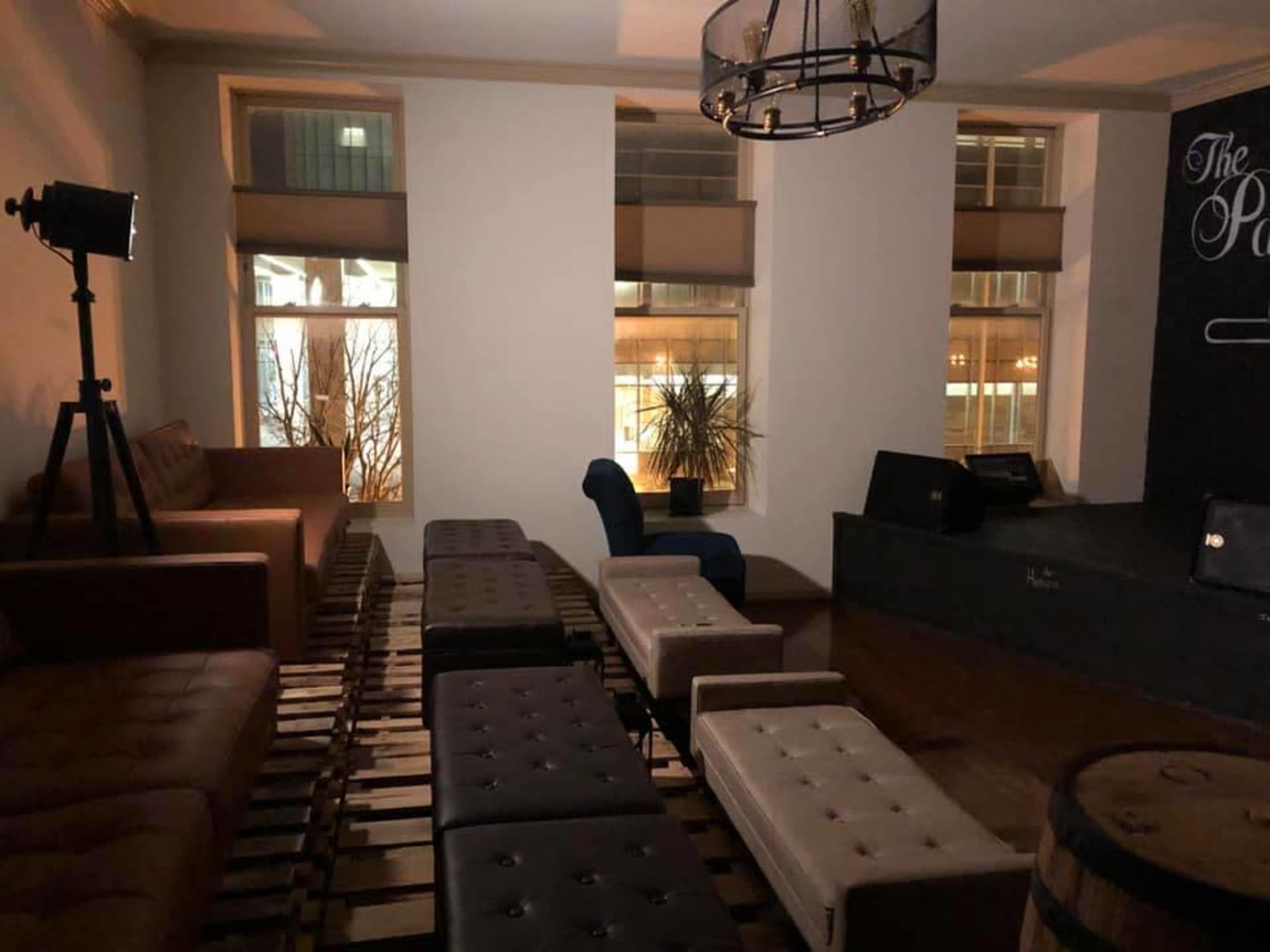 A cozy lounge area with a mix of seating, including a brown leather couch, white ottomans, and a blue chair, illuminated by soft lighting from windows.