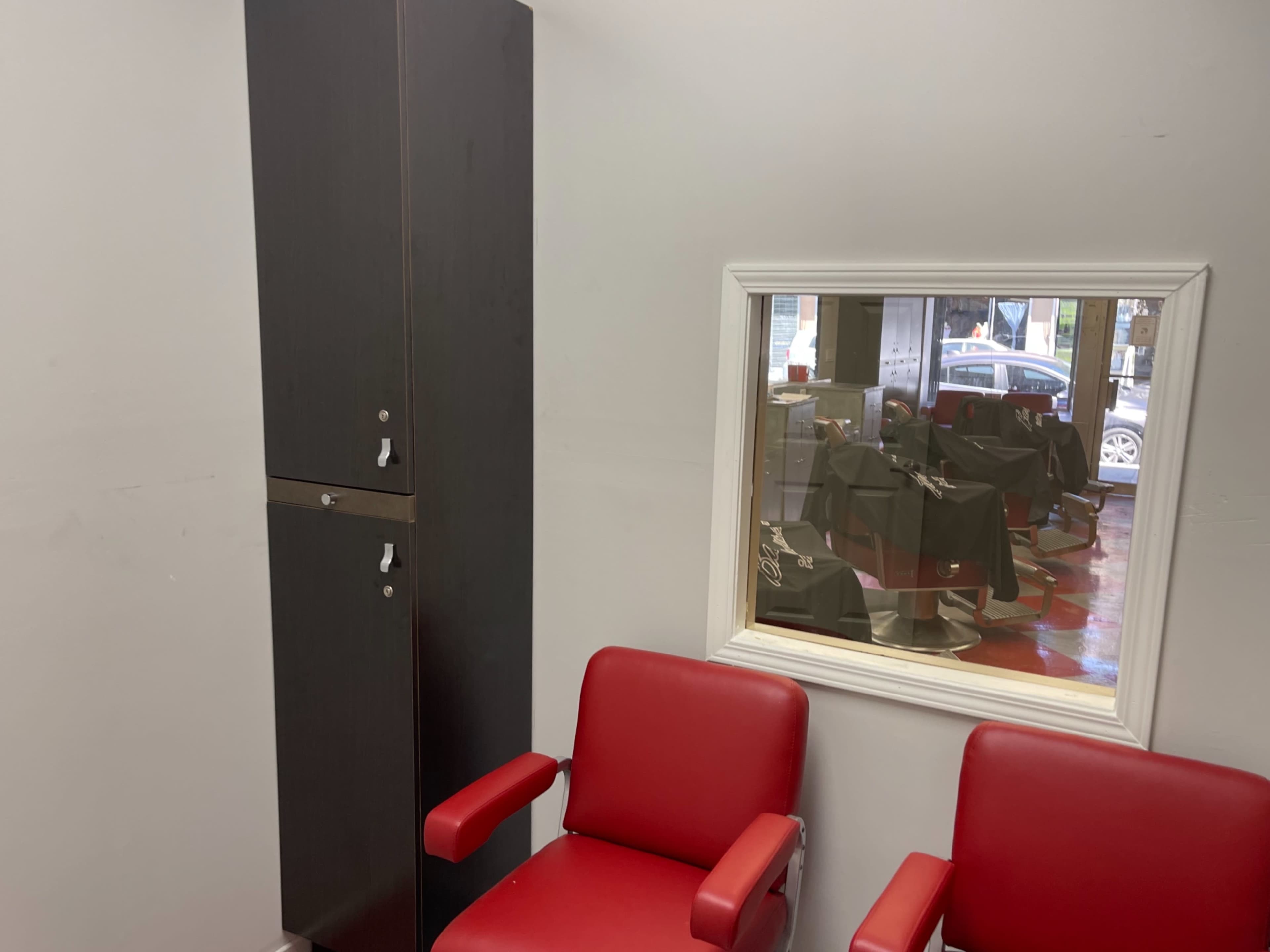 The image shows a pair of red chairs positioned in front of a wall-mounted mirror, with a storage cabinet next to them and a view of barber stations reflected in the mirror.