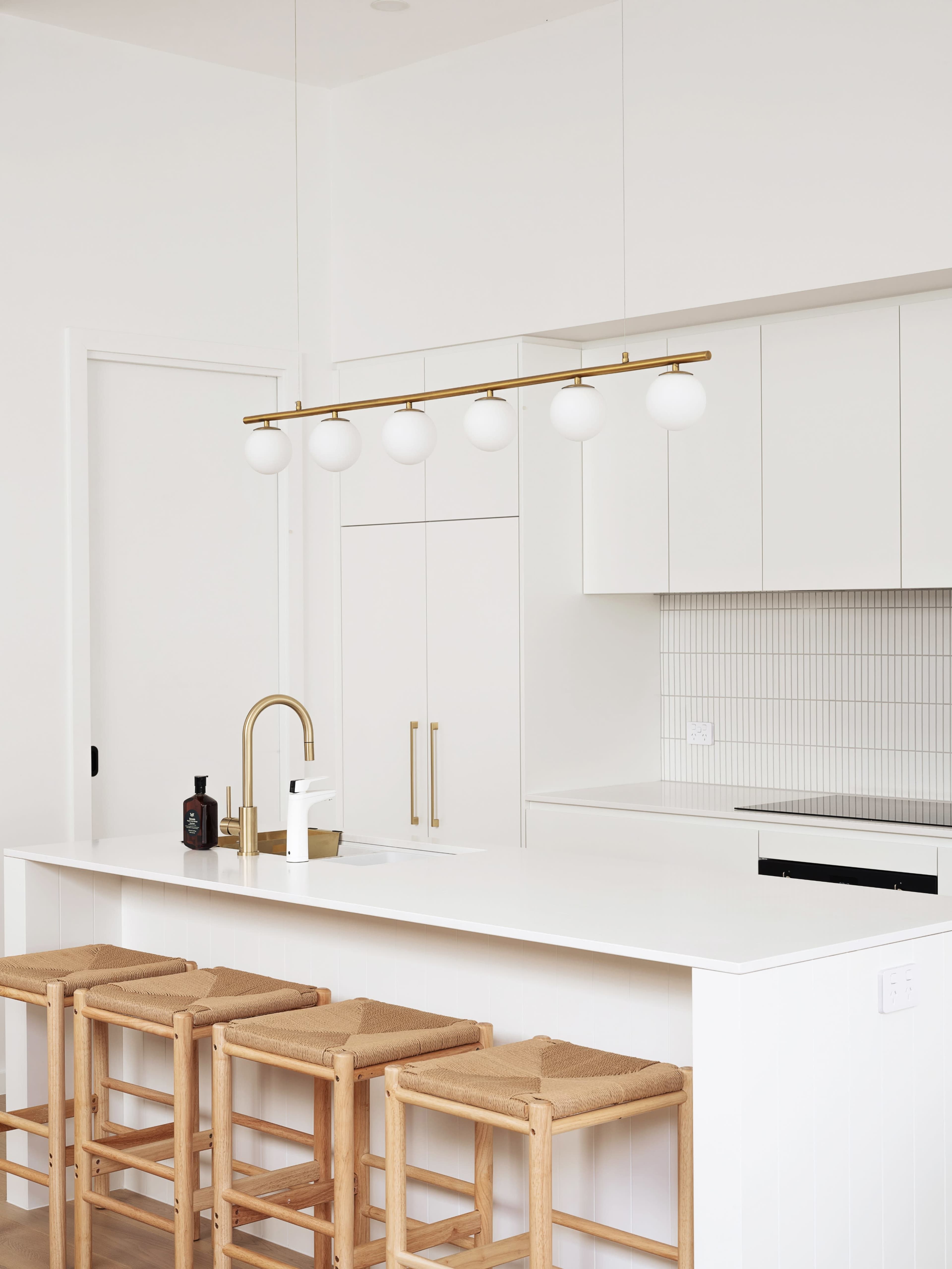 A modern kitchen features a white countertop with three wooden stools, a brass faucet, and a light fixture with five spherical lights hanging above.