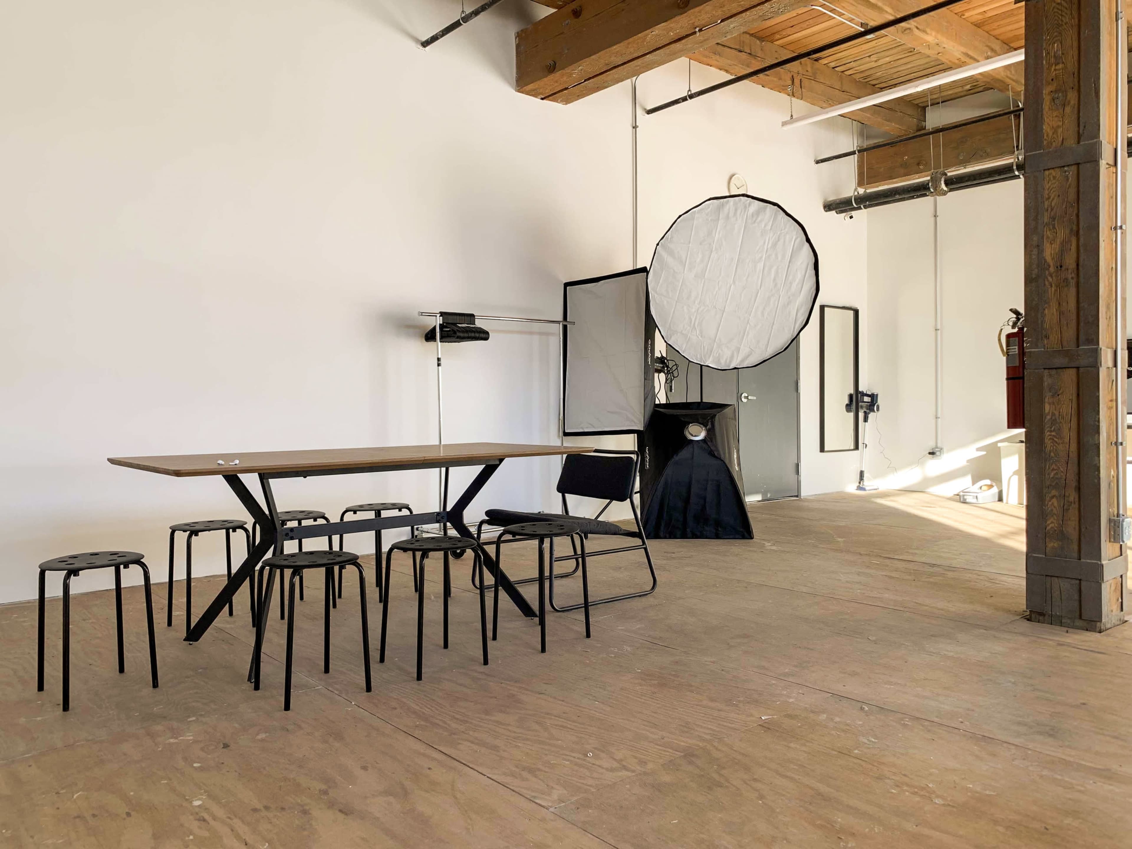 A minimalist studio space features a wooden table surrounded by black chairs, with a lighting setup in the background.