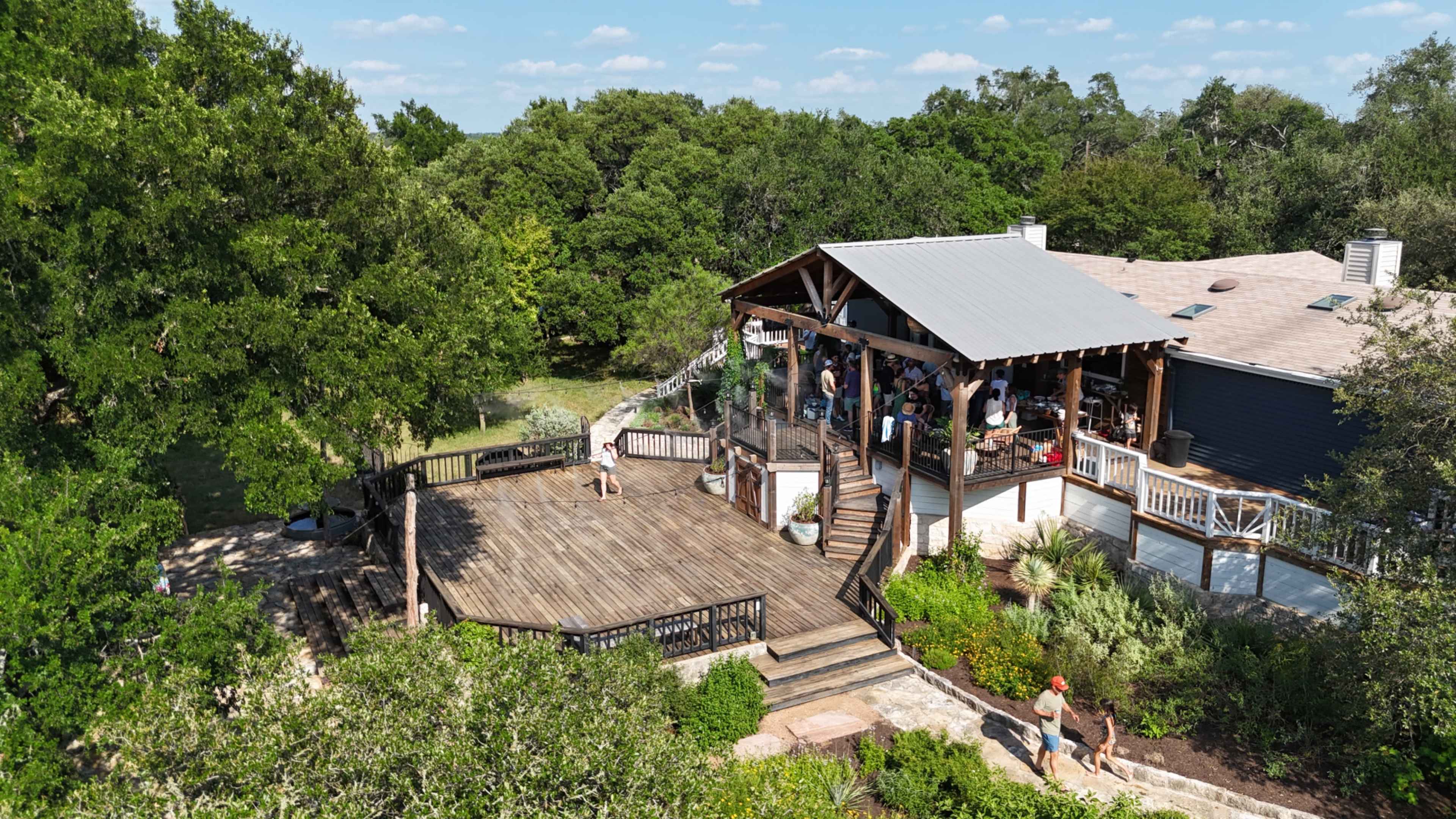 The image shows a large wooden deck attached to a house, surrounded by greenery and a clear blue sky, with several people gathered on the deck and patio areas.