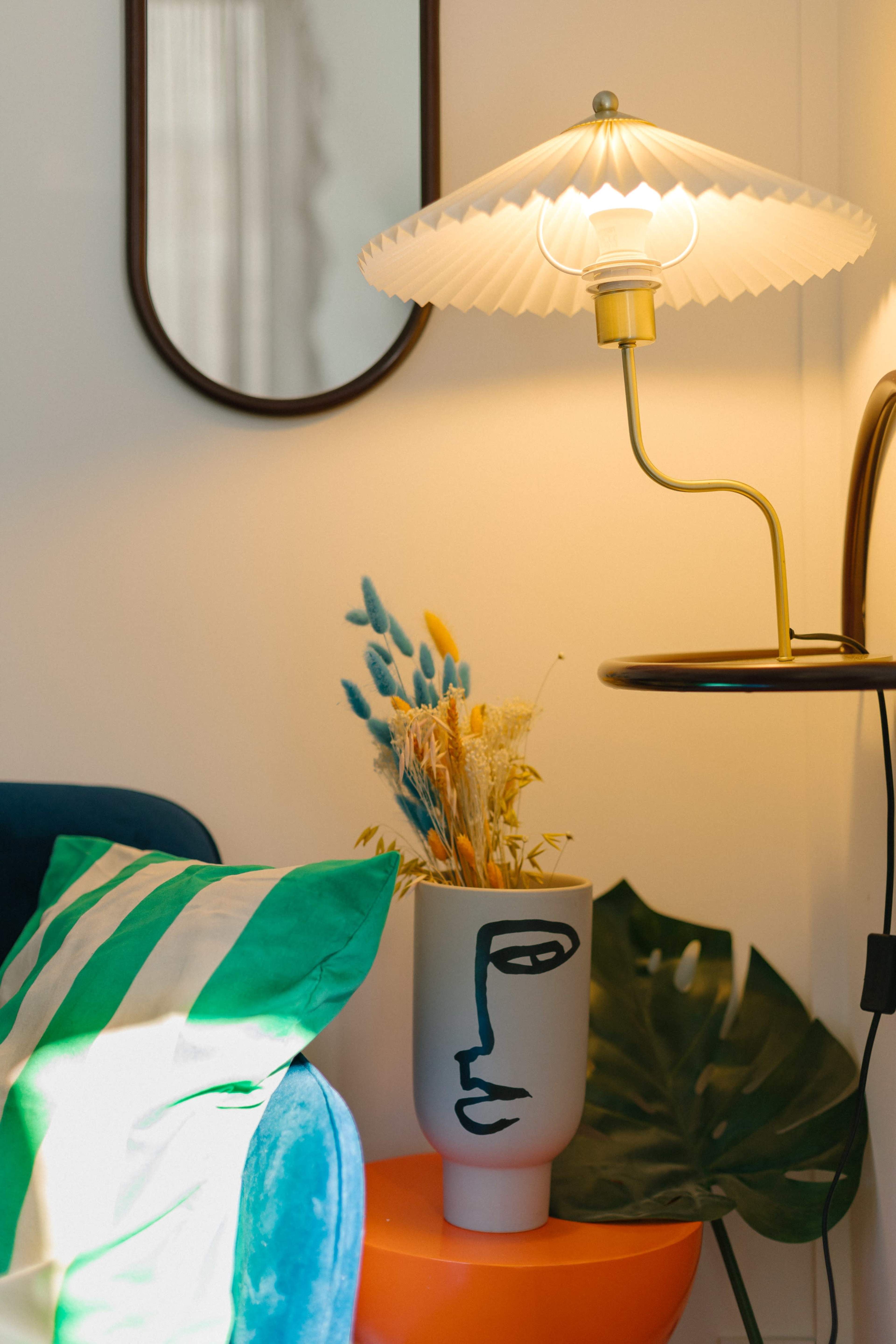 A lamp with a pleated shade stands next to a decorative vase featuring a face design, surrounded by dried flowers and a green and white striped pillow on a blue couch.