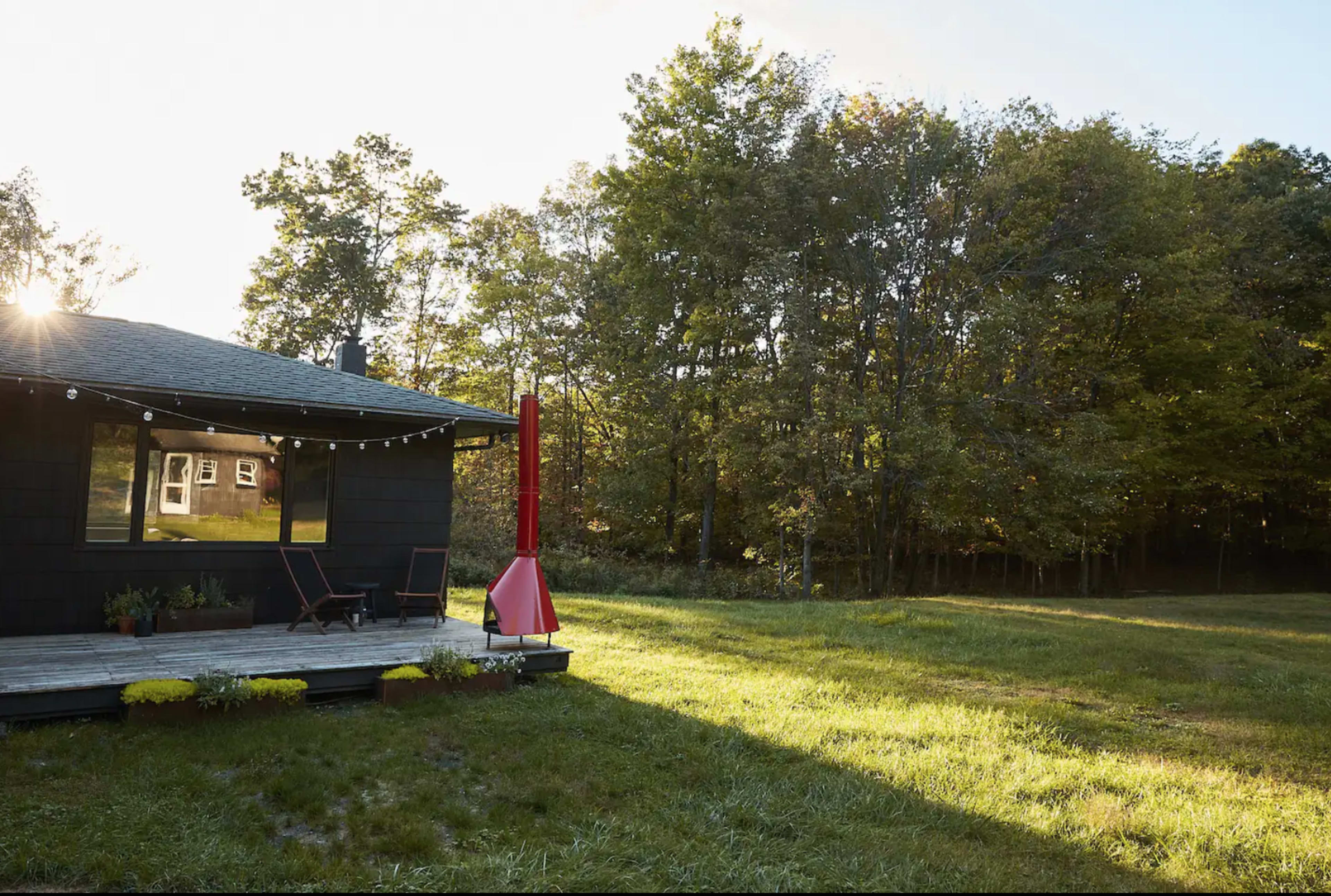 A modern cabin with large windows and a red chimney stands on a wooden deck in a grassy area surrounded by trees.