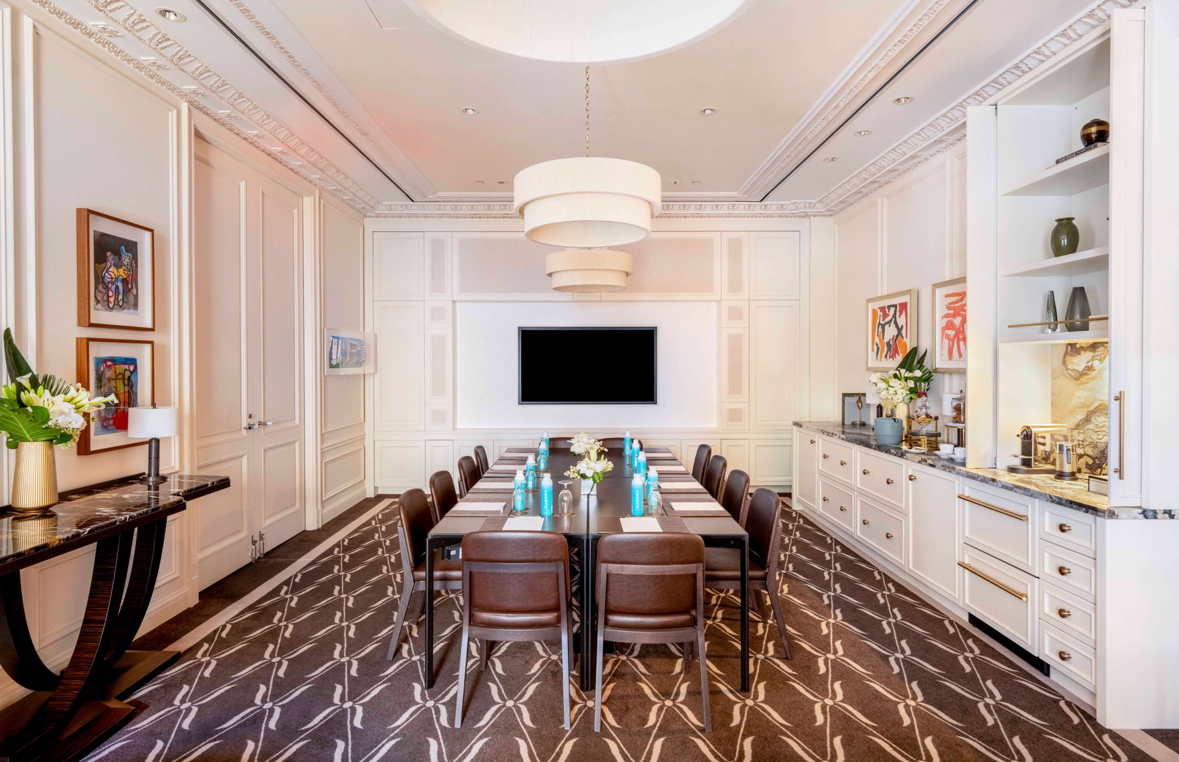 The image shows a modern conference room featuring a long table with ten brown chairs, a large wall-mounted screen, and elegant decor including a chandelier and side cabinet.